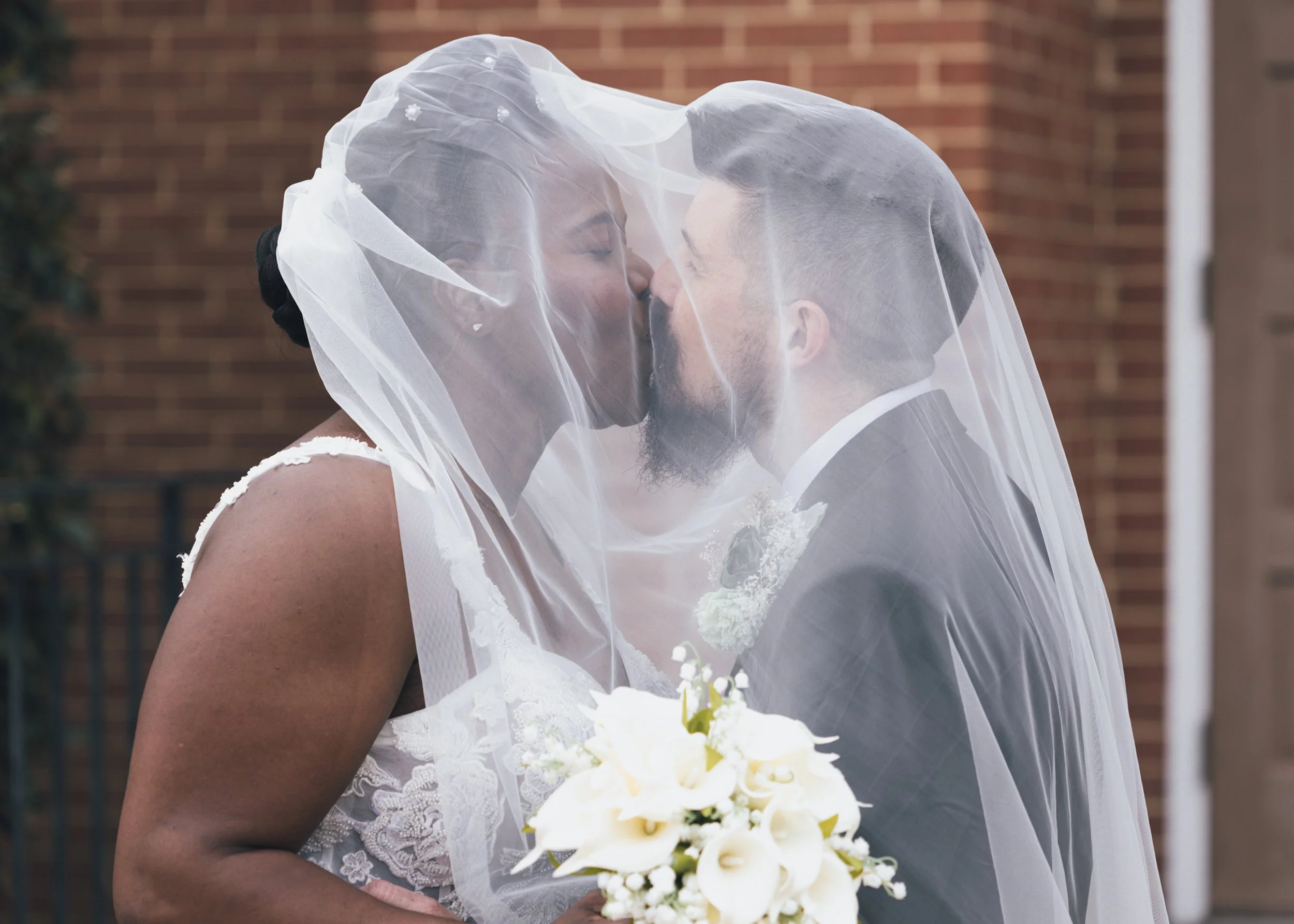 A bride and groom sharing a kiss, with the bride wearing a veil and holding a bouquet of white flowers, standing outdoors in front of a brick wall.