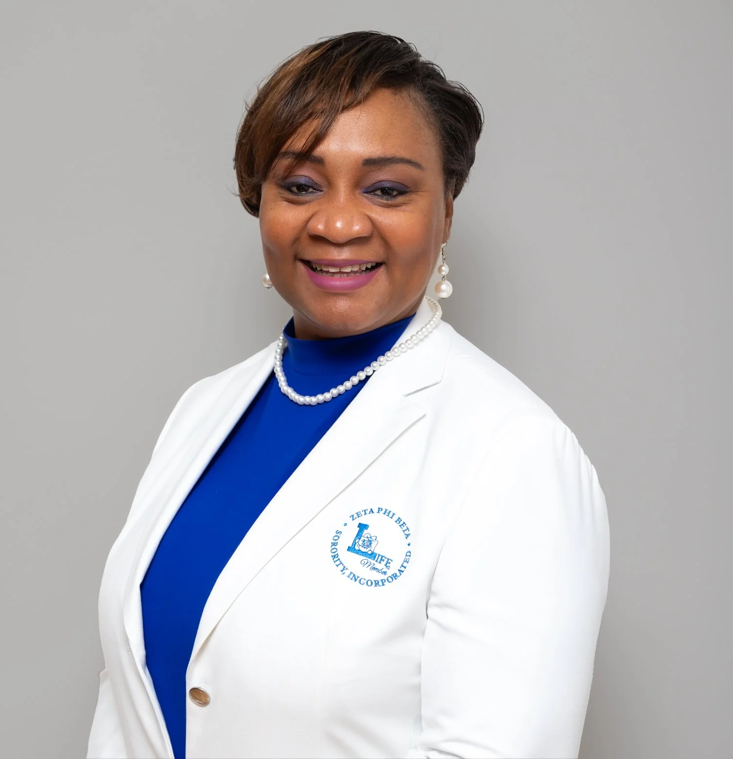 A professional woman with short brown hair, wearing a white blazer with a Zeta Phi Beta sorority emblem, a blue top, pearl earrings, and a pearl necklace, smiling against a plain gray background.