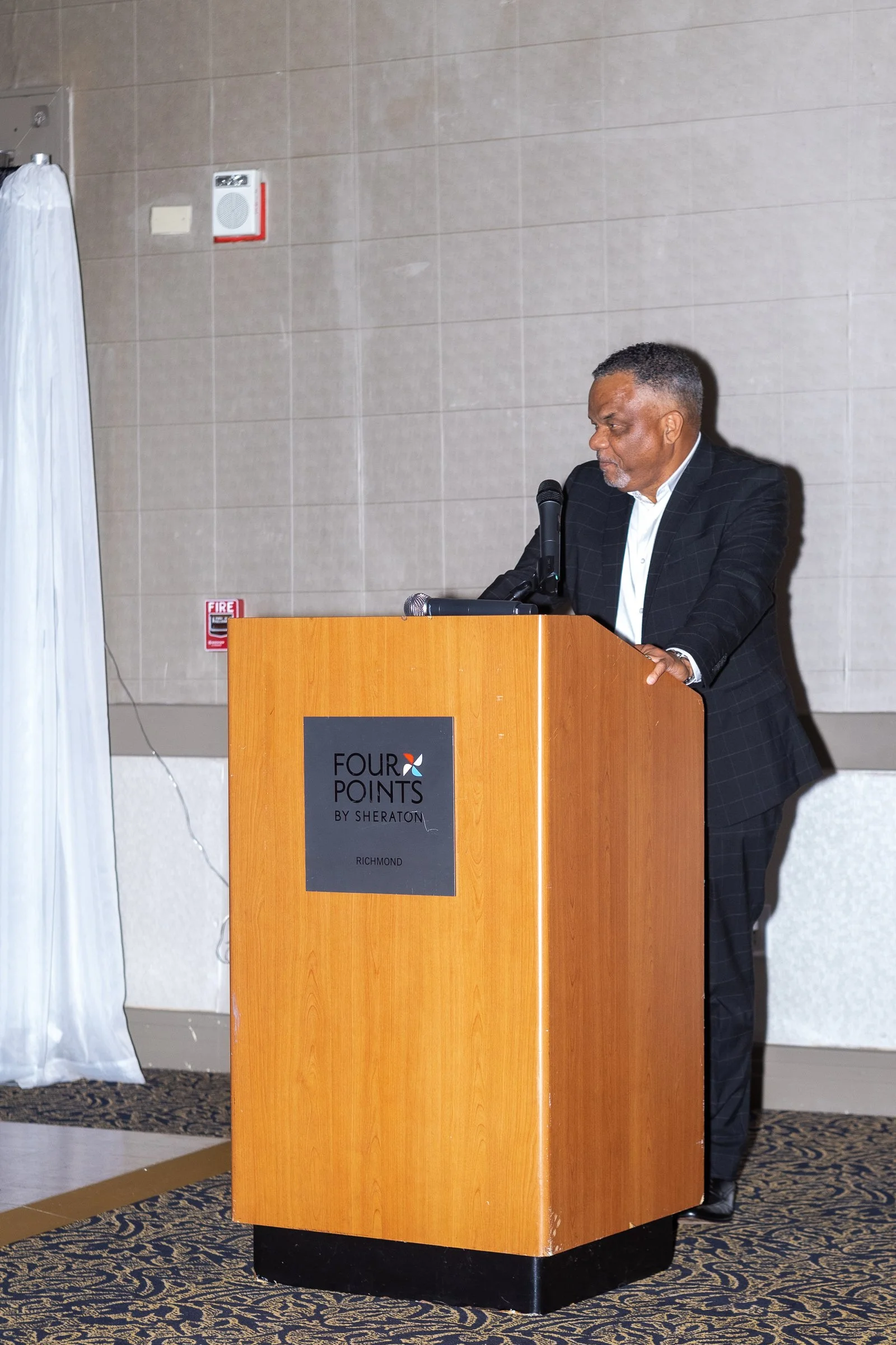 A man with gray hair, dressed in a black checkered suit and white shirt, is standing at a podium with the logo of Four Points by Sheraton, Richmond, speaking into a microphone.