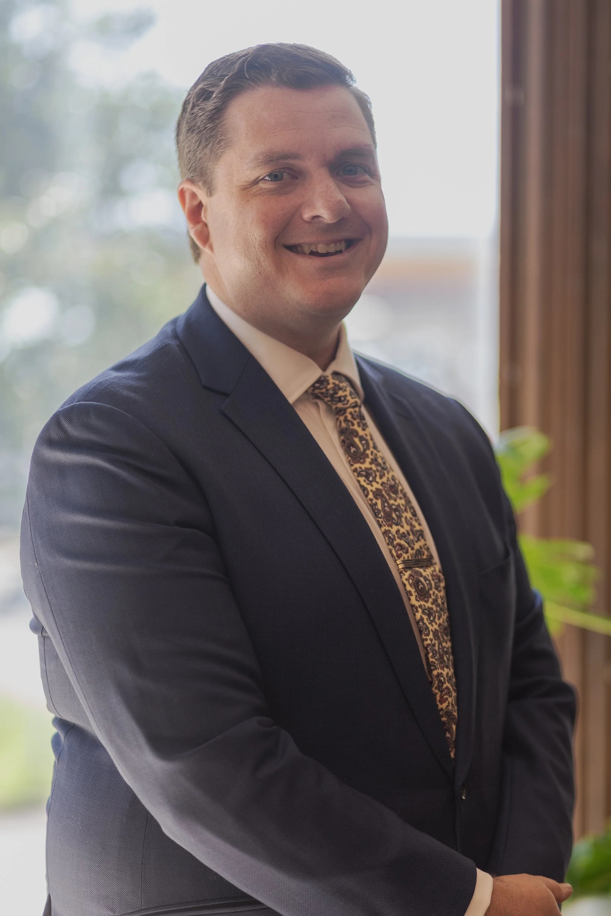 A man in a navy blue suit, white shirt, and floral tie smiling, standing indoors near a window with a blurred outdoor scene in the background.