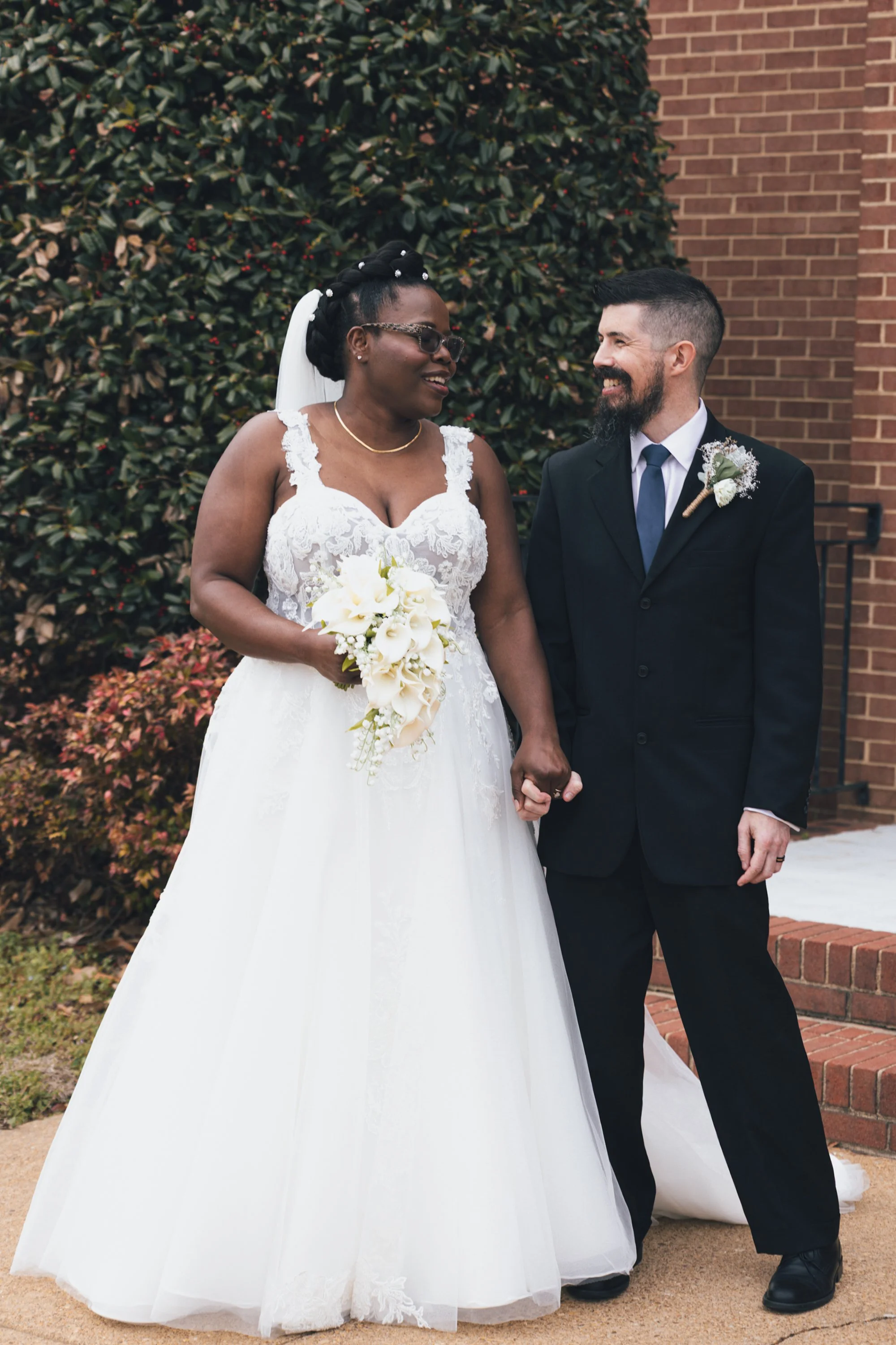 A bride and groom smiling and holding hands outdoors in front of a brick wall and green shrubbery on a wedding day.