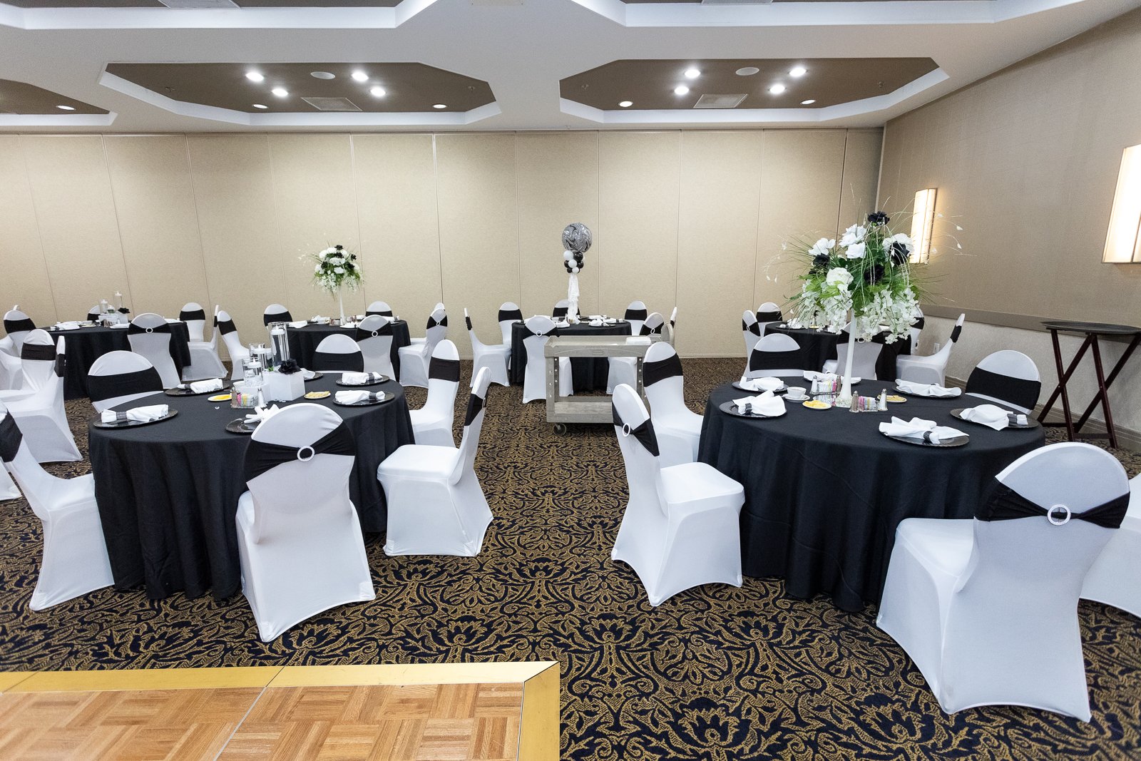Banquet hall decorated for an event with round tables covered in black tablecloths and white chair covers with black sashes. Tall floral centerpieces with white flowers and greenery are on the tables. The ceiling has recessed lighting and the floor h