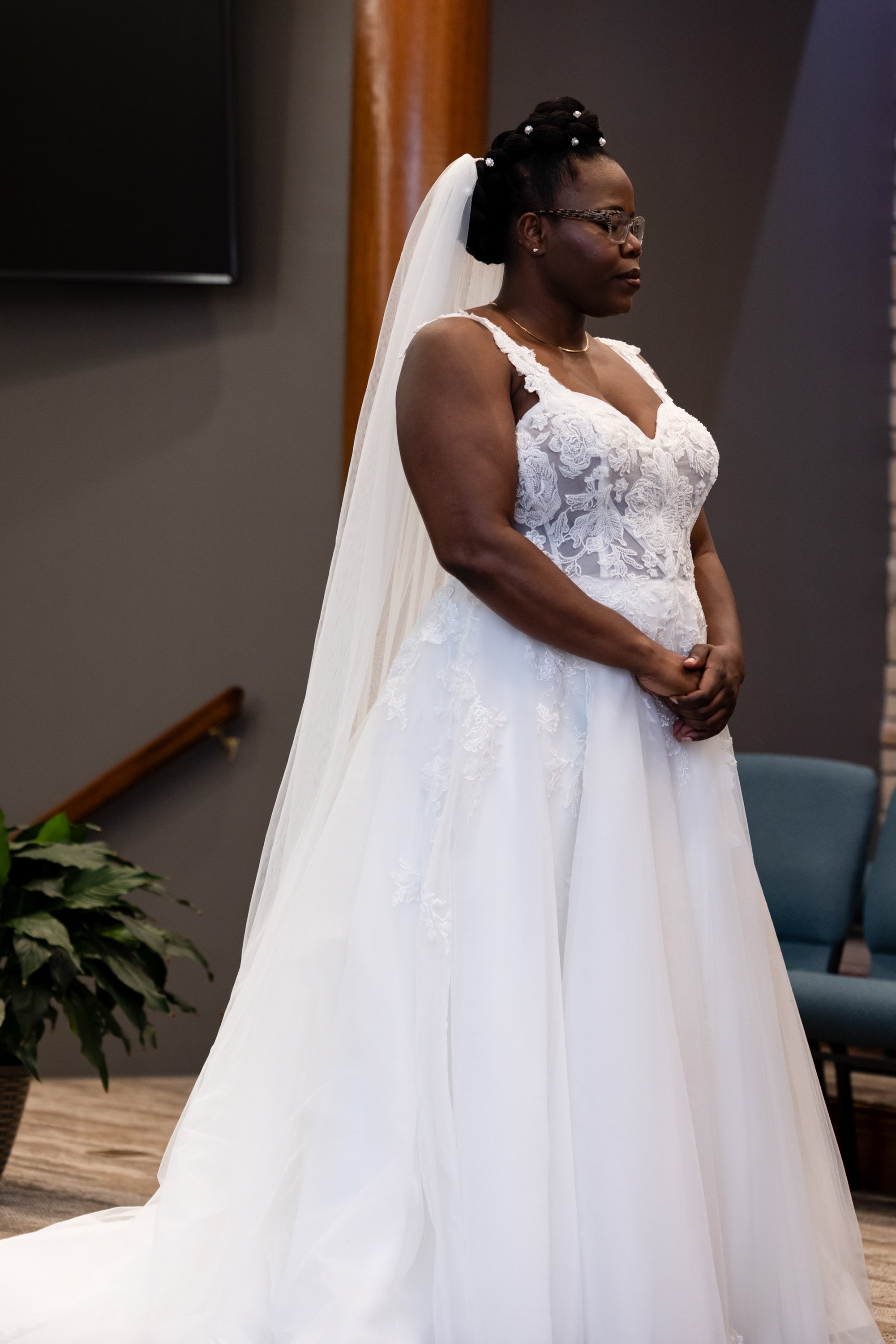 A woman wearing a wedding dress with lace details and a veil, standing with her eyes closed and hands clasped in front of her.