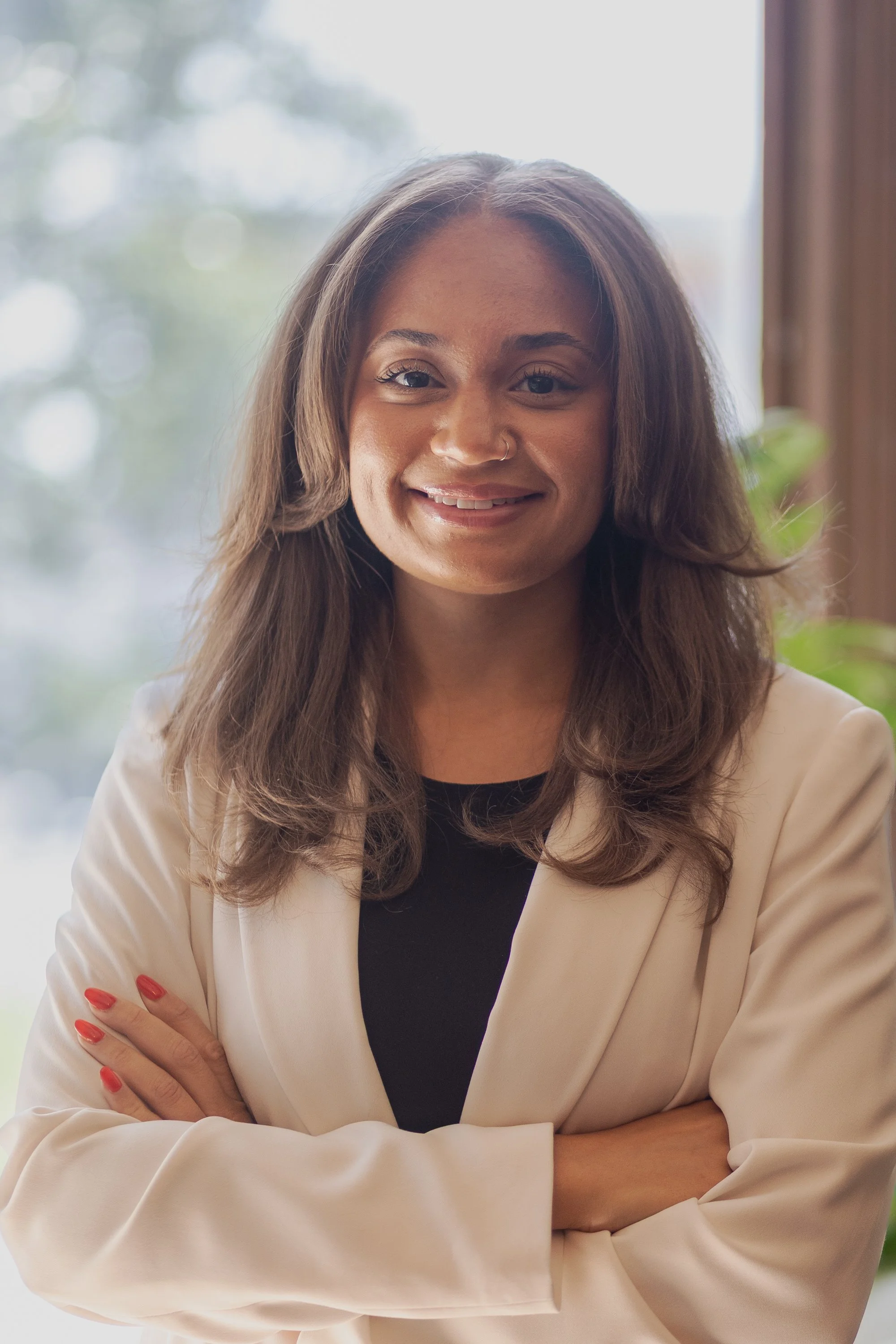 A woman with long brown hair, wearing a beige blazer over a black top, smiling with arms crossed in front of a softly blurred background with natural light.