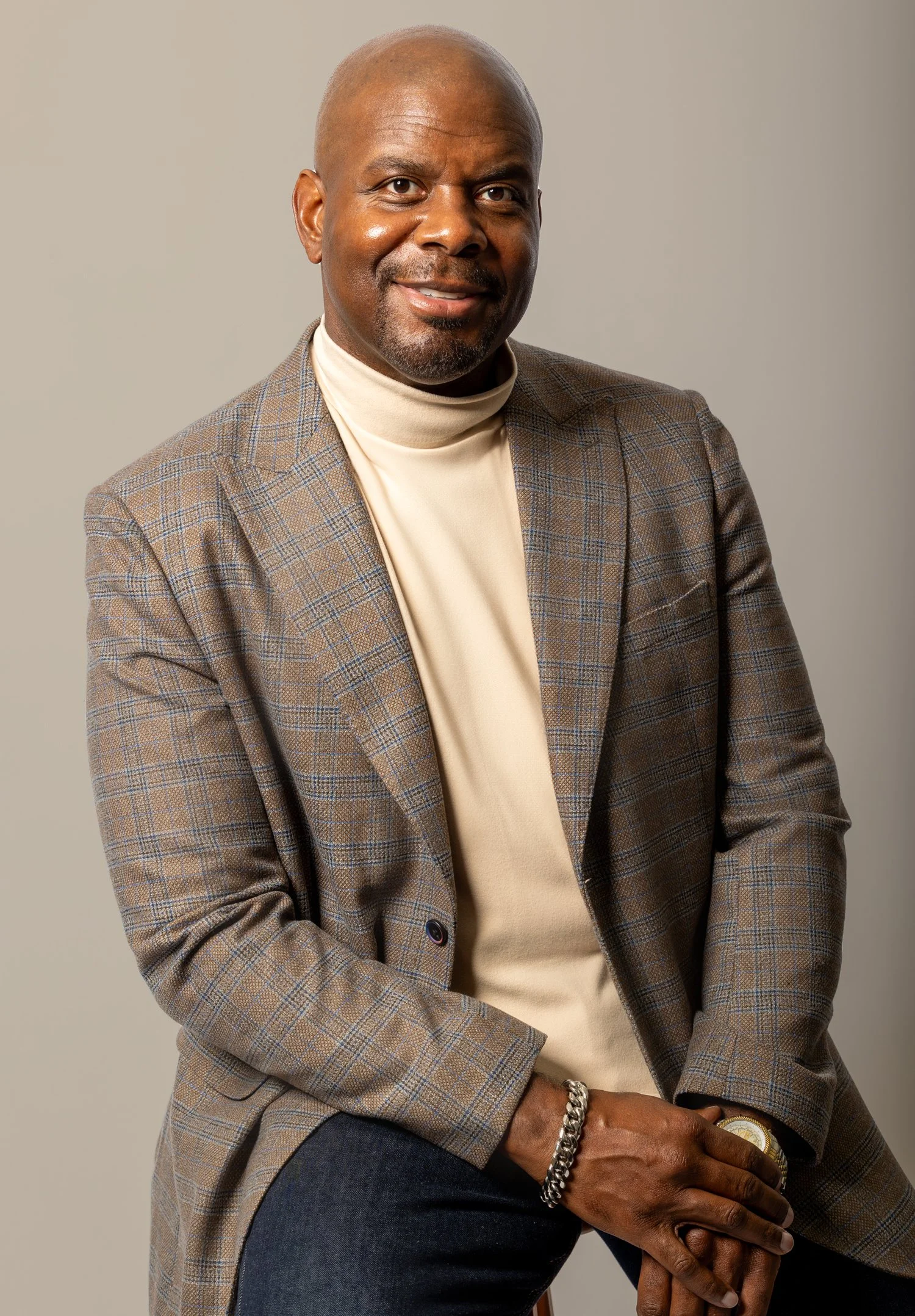 A middle-aged man with a shaved head and goatee, wearing a beige turtleneck sweater, a brown plaid blazer, dark jeans, a gold watch, and a bracelet, sitting against a plain beige background.
