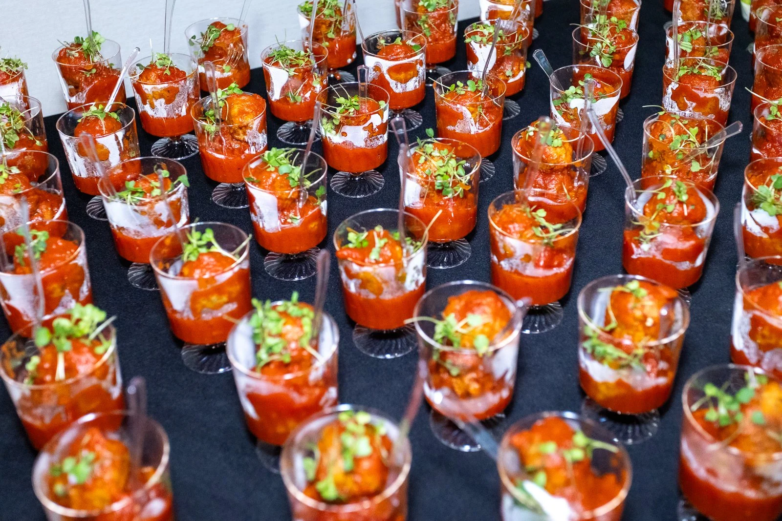 Multiple small glasses filled with tomato-based appetizers garnished with microgreens, arranged on a black surface at a catering event.