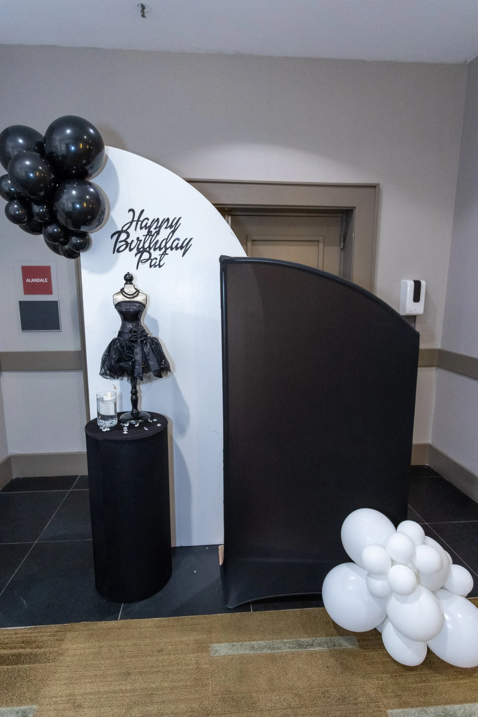 Backdrops with black balloons, a dress form, and a cluster of white balloons, set up for a birthday celebration.
