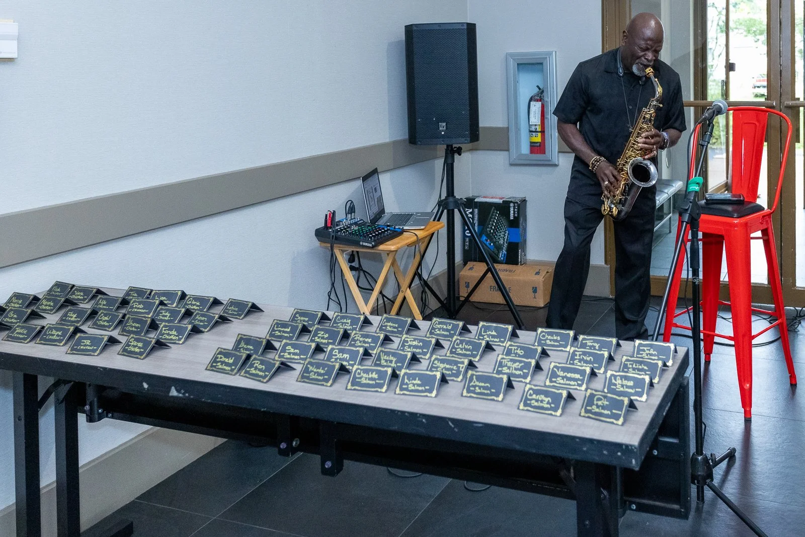 A man playing saxophone at a social event with name cards laid out on a table in front of him, sound equipment, and a microphone stand nearby.