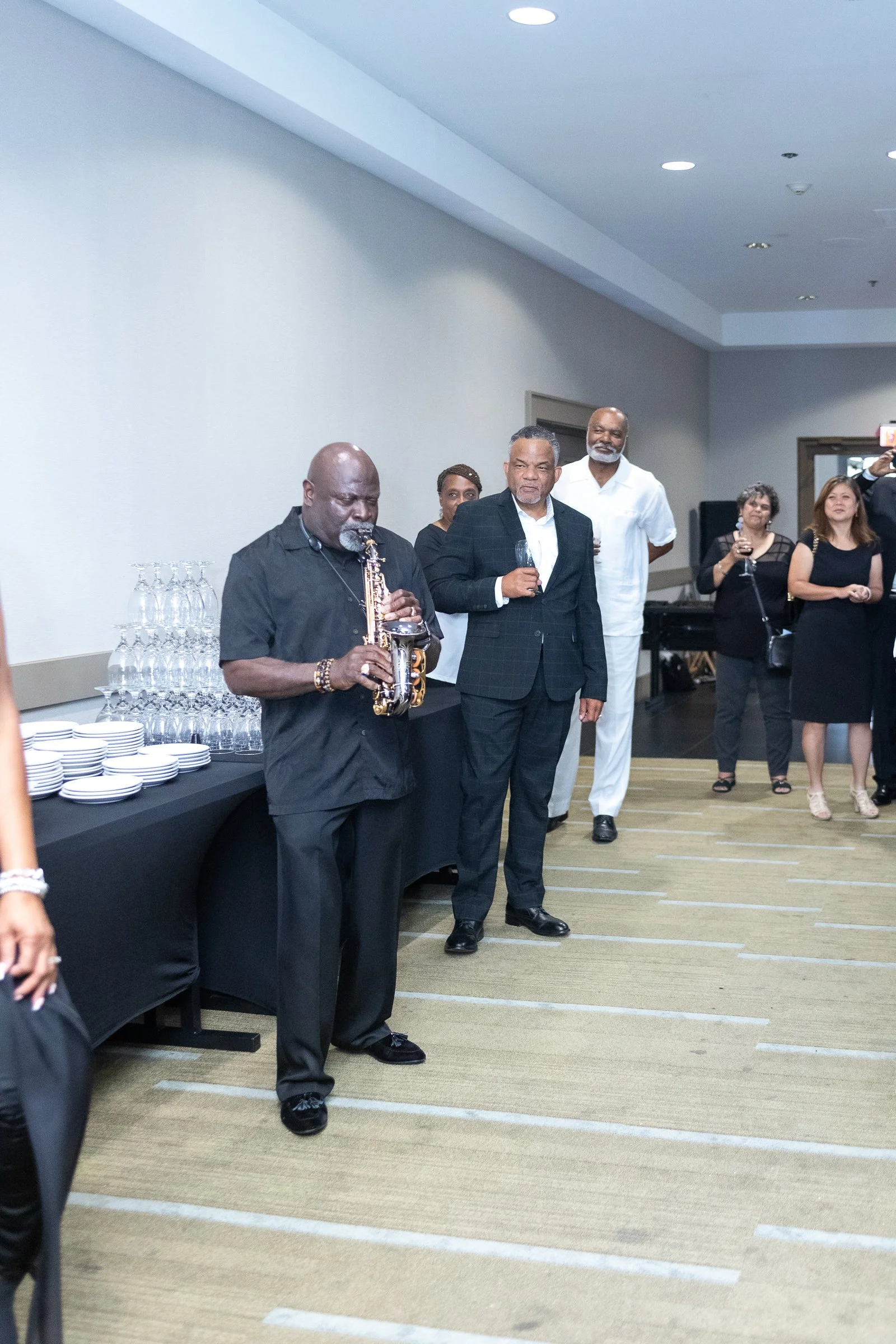 A group of people at a social event, with a man playing saxophone and others holding wine glasses, standing in a room with a refreshment table on the left.