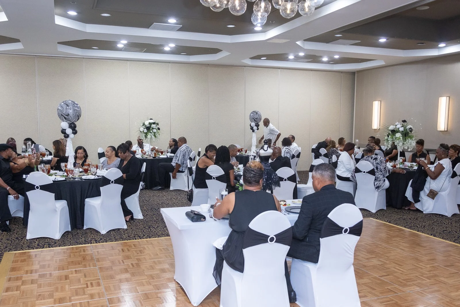 People seated at round tables with black and white chairs at a formal event in a banquet hall decorated with black and white balloons and floral centerpieces.