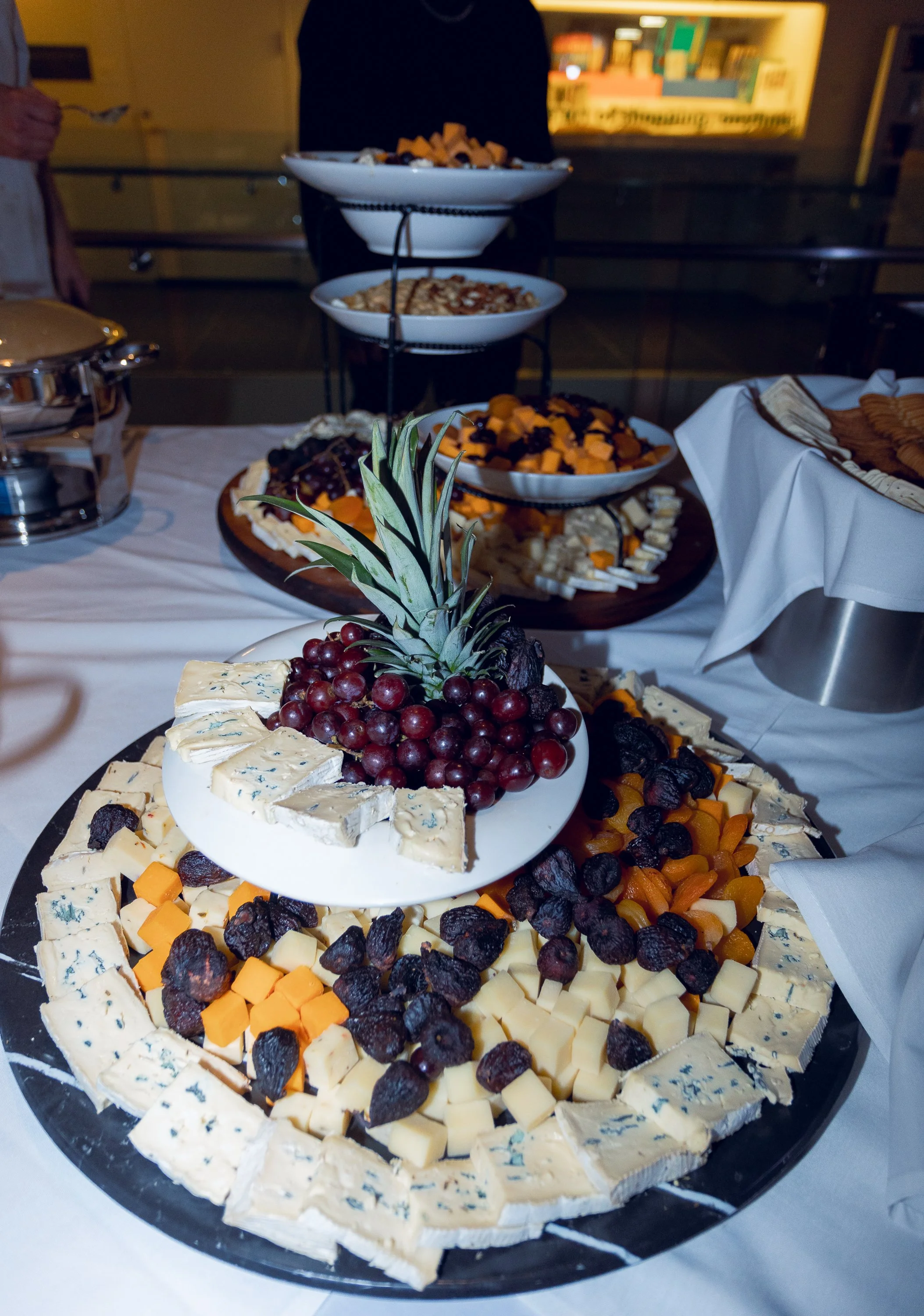 A cheese and fruit platter with a pineapple decoration centerpiece, featuring various types of cheese, dried fruits, and fresh grapes, surrounded by more cheese and fruit on a black tray.