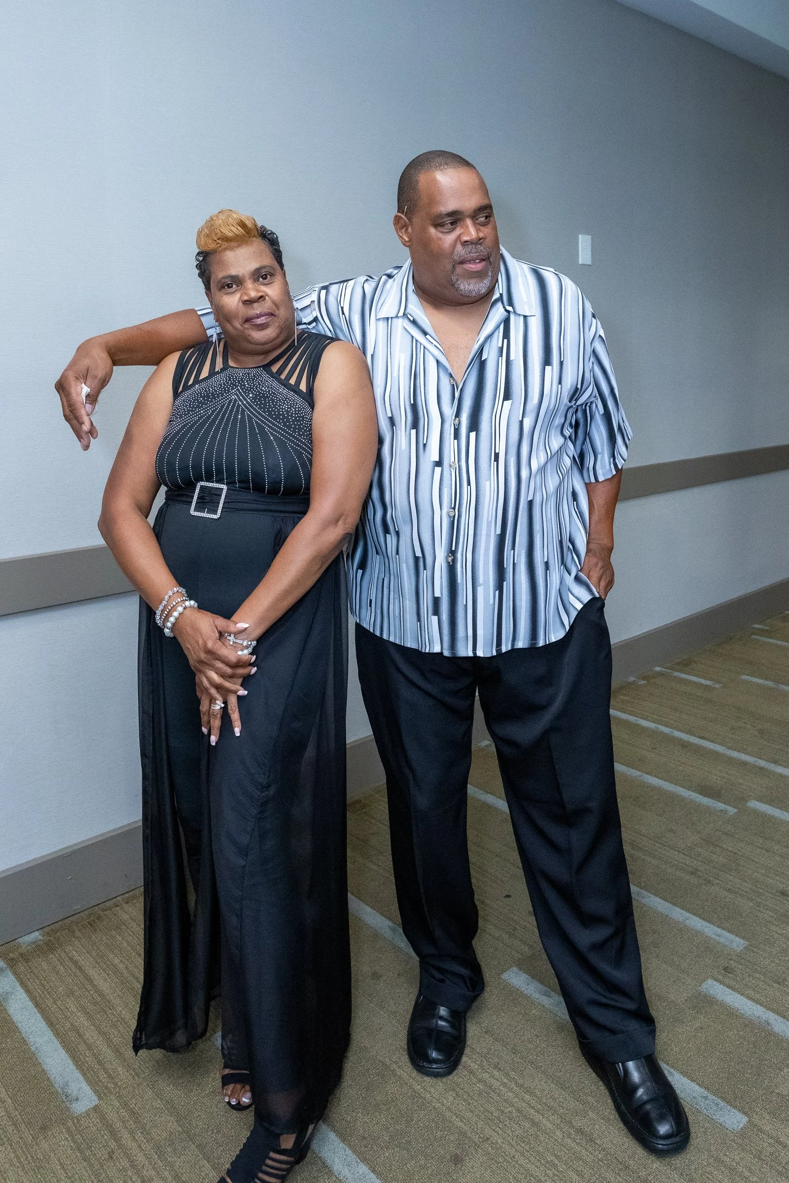A man and a woman standing close together in a hallway, with the man placing his arm around the woman's shoulder. They are dressed in casual to semi-formal clothing, with the woman wearing a black dress and the man wearing a striped shirt and black p
