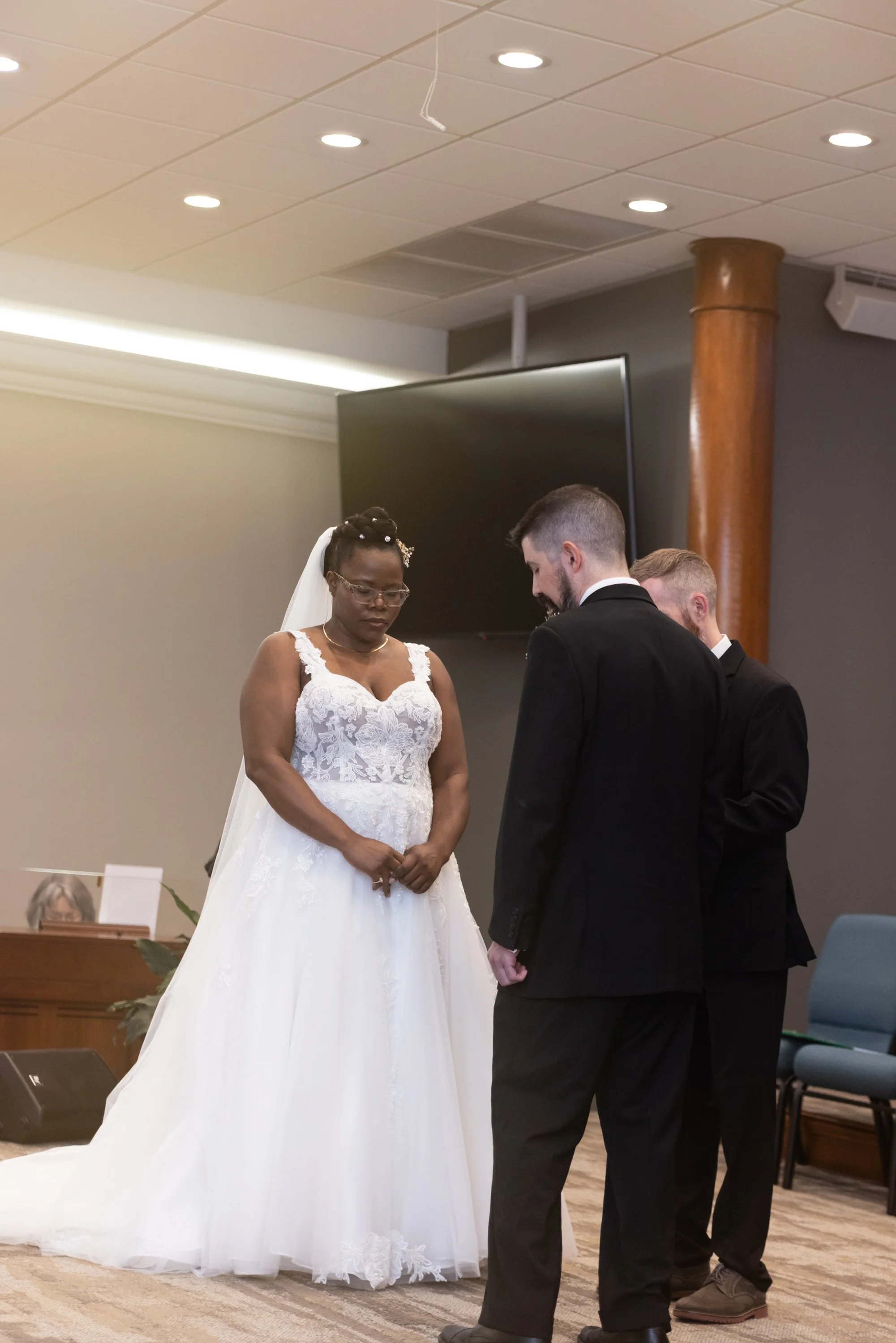 A couple getting married during a wedding ceremony with the officiant or priest present. The bride is wearing a white wedding gown with lace details and a veil, and the groom is dressed in a dark suit. They are holding hands and standing in front of 