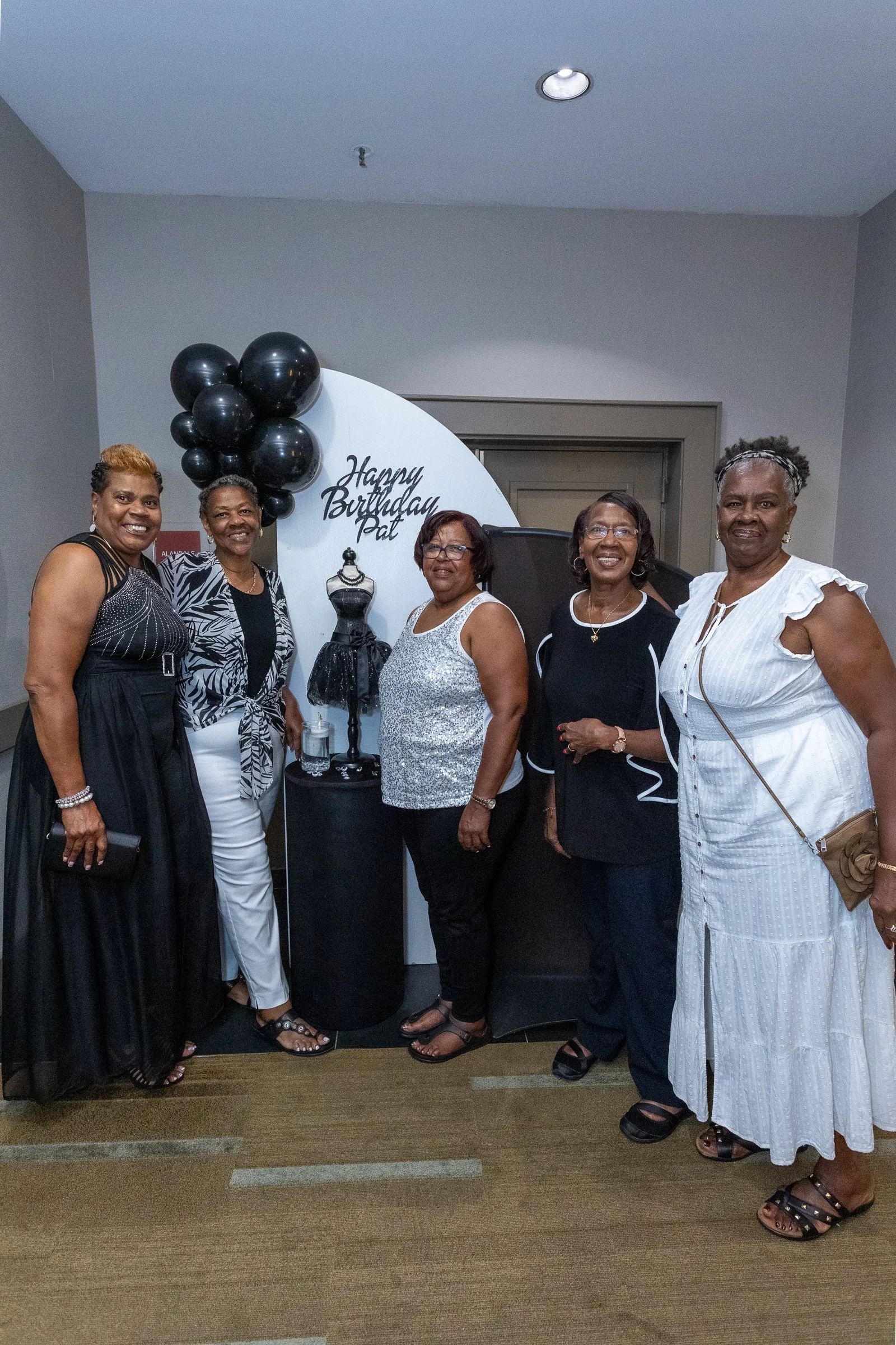 Five women standing in front of a white semi-circular backdrop with black balloons and a dress mannequin, celebrating a birthday, with the words 'Happy Birthday Pat' written on the backdrop.