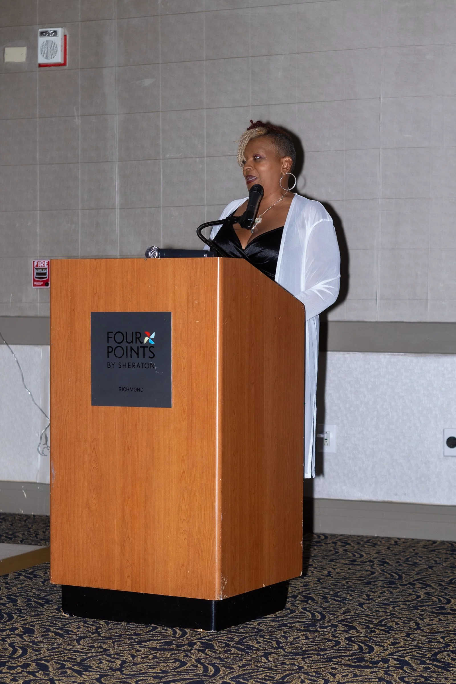 Woman speaking at a podium in a conference room at Four Points by Sheraton Richmond hotel. The woman has short, styled hair and is wearing hoop earrings, a black top, and a white cover-up. The podium is wooden with a black base, and the floor has a p