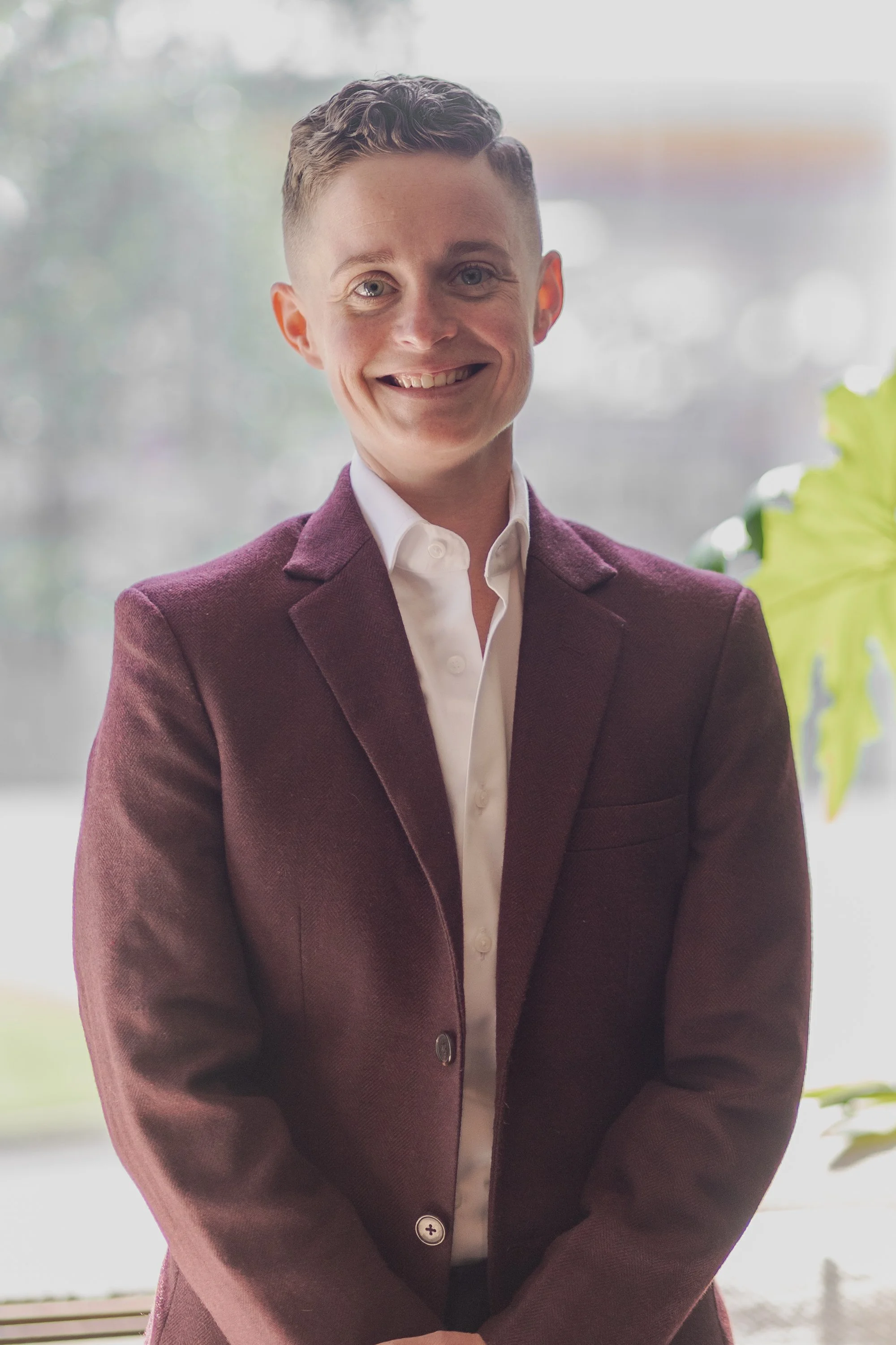 A young man with short curly hair, wearing a white shirt and a maroon blazer, smiling with his arms crossed, standing indoors near a window with a blurred outdoor background.
