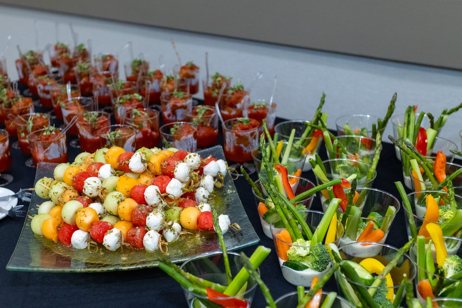 Assorted appetizers including cherry tomato and mozzarella skewers, vegetable sticks in small cups, and mini beef tacos with cilantro serve at a catering event.