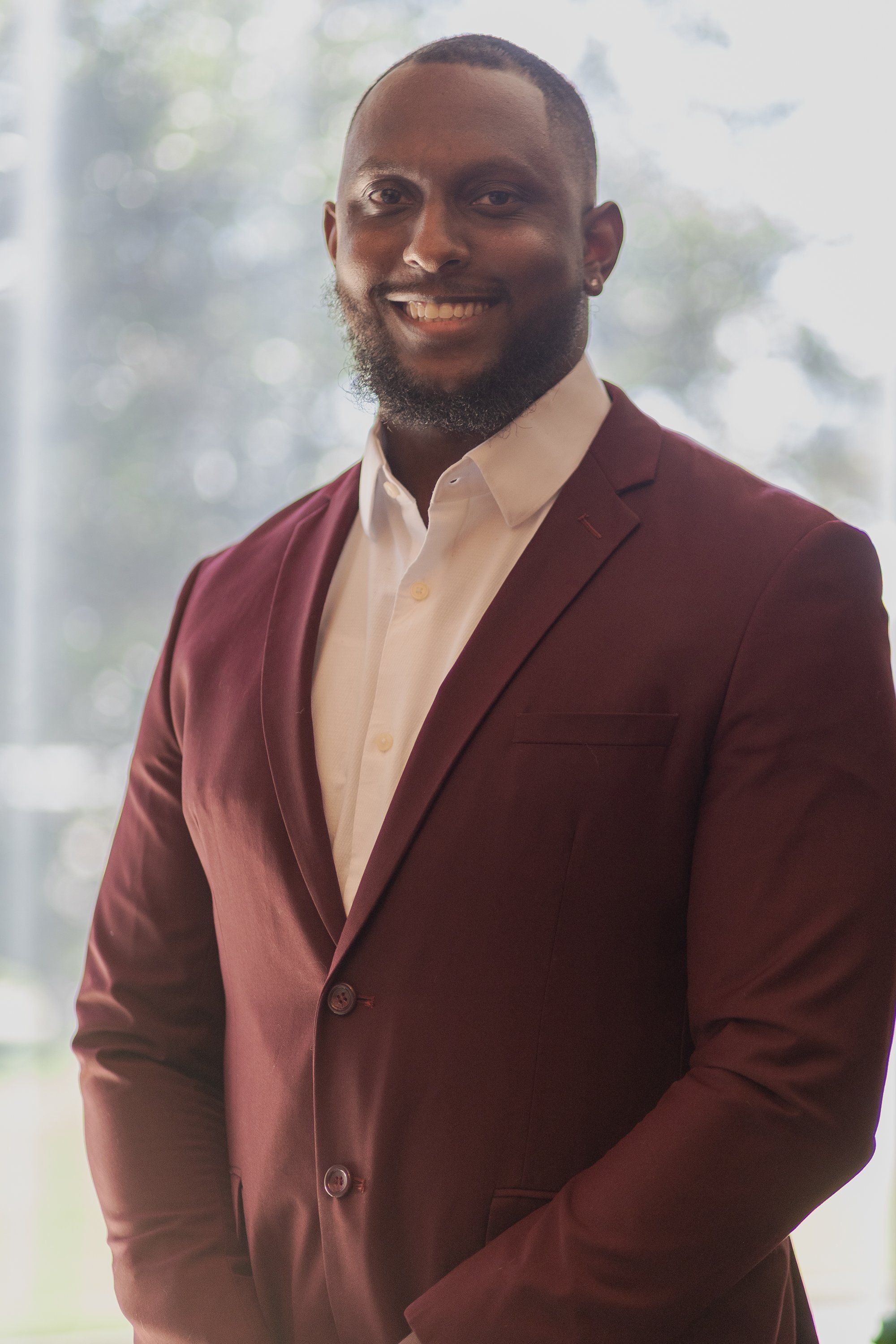 A man with short hair and a beard smiling, wearing a maroon suit jacket and a light-colored shirt, standing indoors with a blurred background of trees and light.
