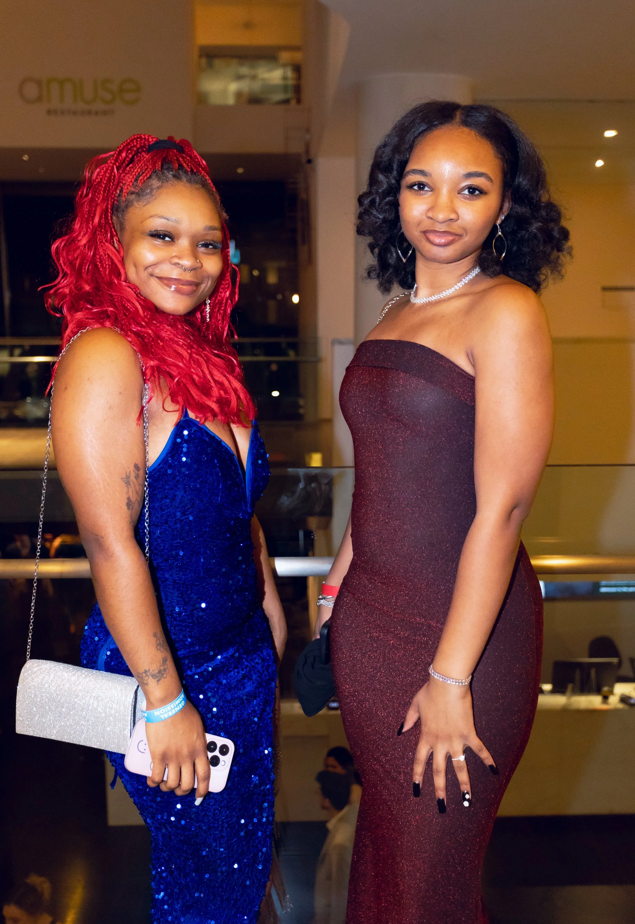 Two young women dressed in elegant evening gowns, one in a blue sequined dress and the other in a burgundy dress, posing at a formal event.
