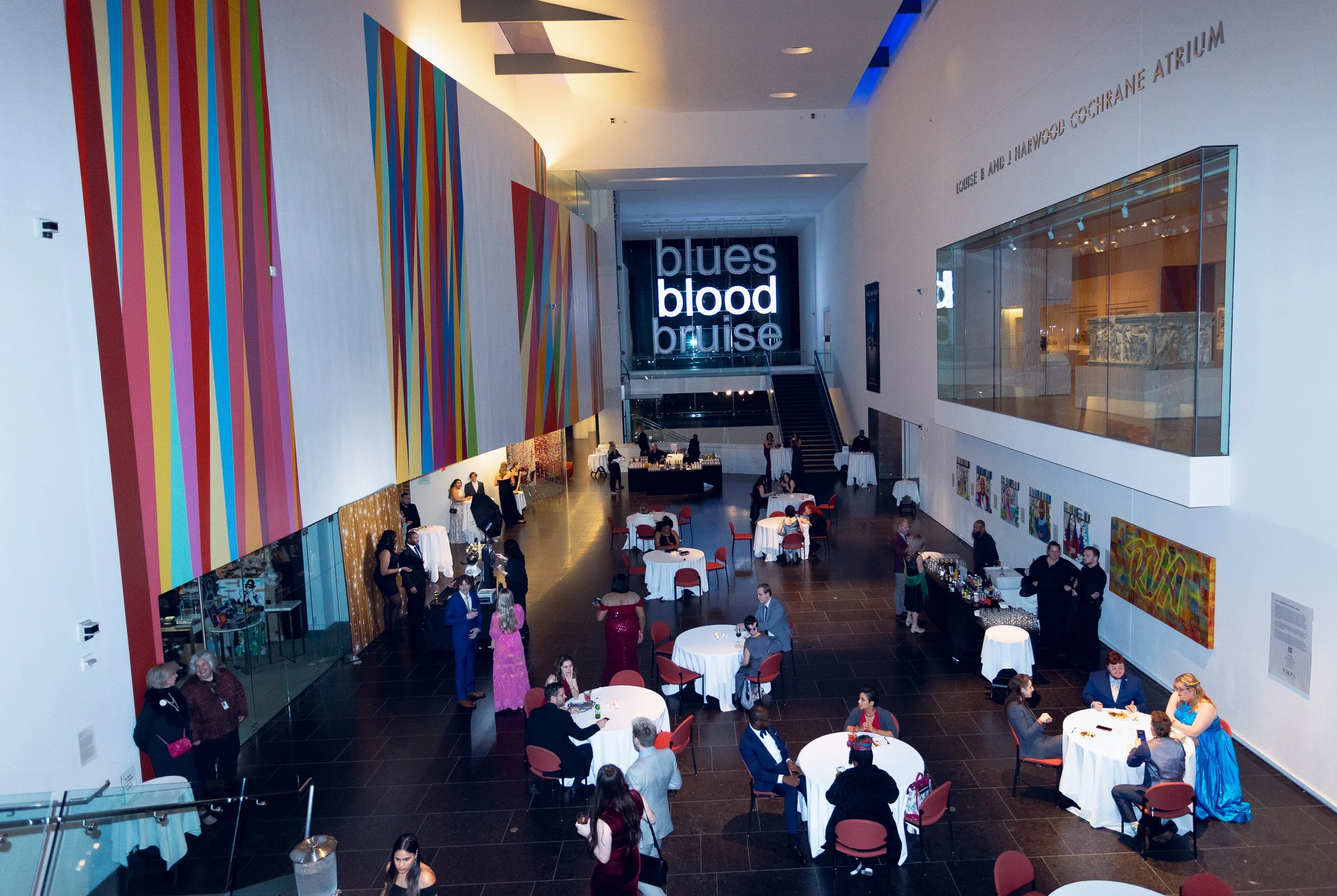 The interior of a modern, multi-story building with a spacious lobby filled with round tables and red chairs. People are mingling, walking, and sitting at the tables. There is colorful, striped wall art on the left, and a large illuminated sign that 