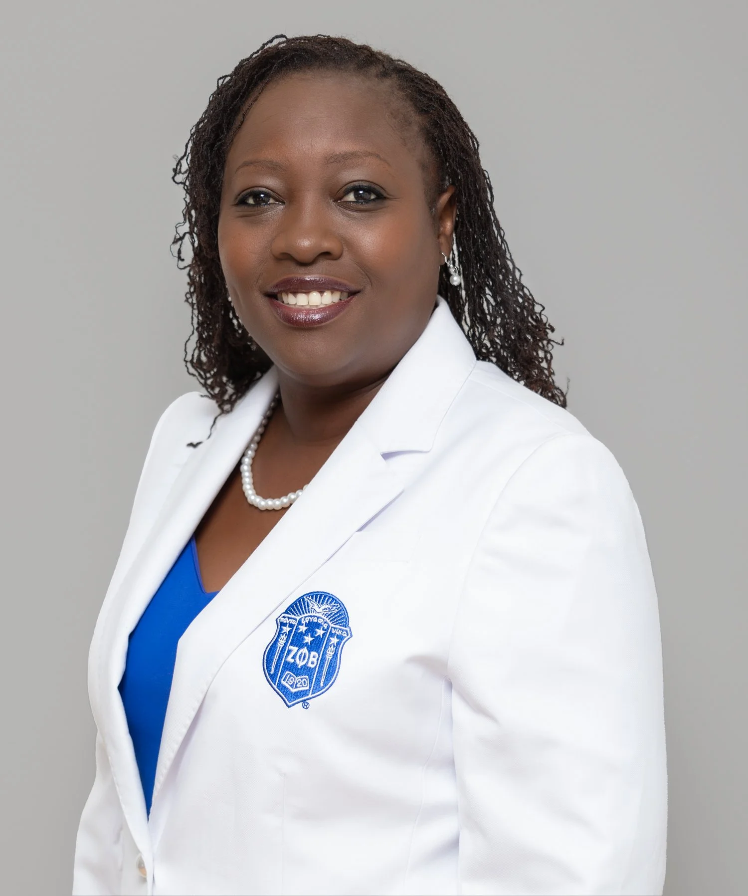 A woman with short curly hair smiling, wearing a white blazer with a blue emblem, pearl earrings, and a pearl necklace, against a plain gray background.