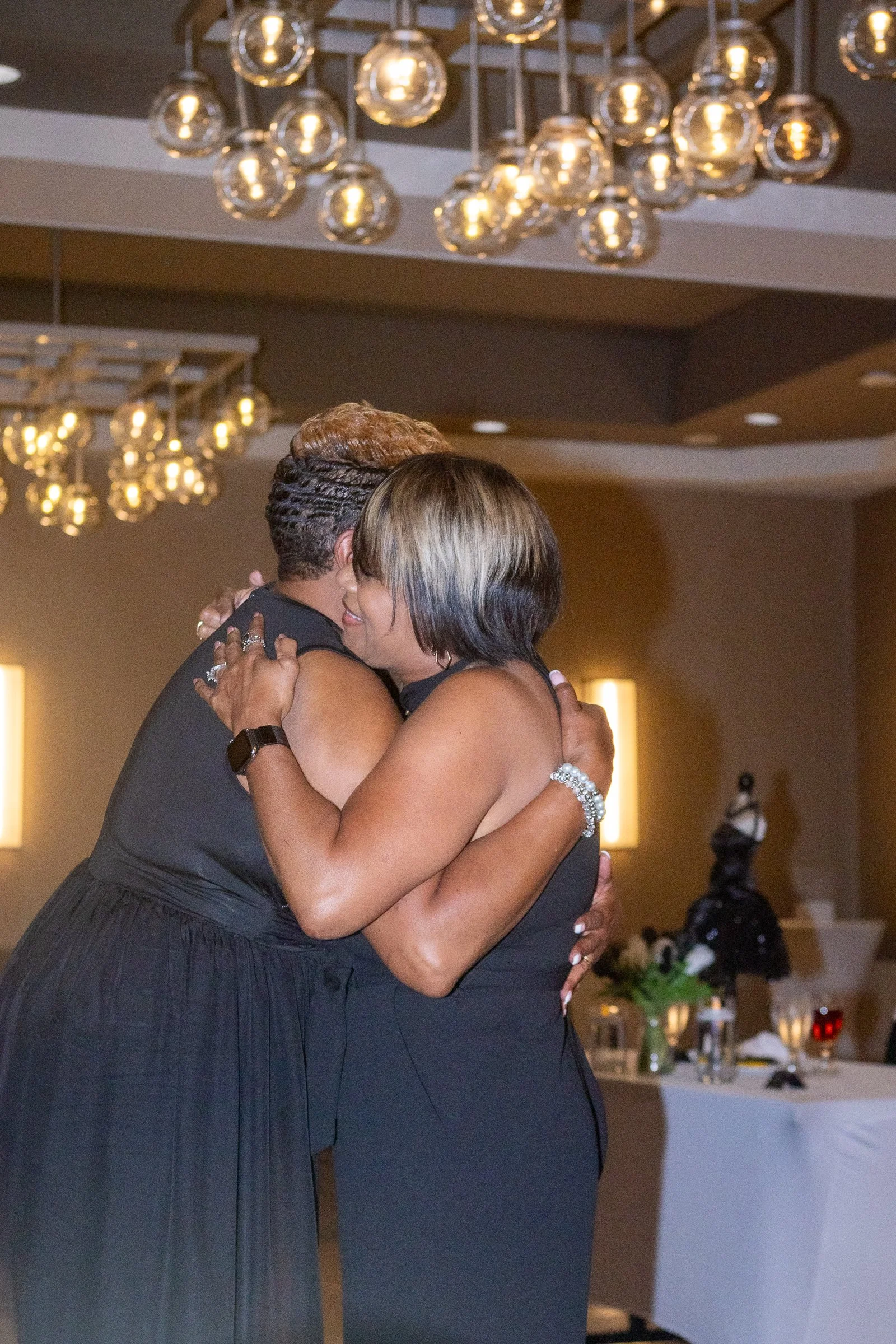 Two women dancing and hugging at a celebration or event in a warmly lit room with hanging light fixtures and a decorated table in the background.