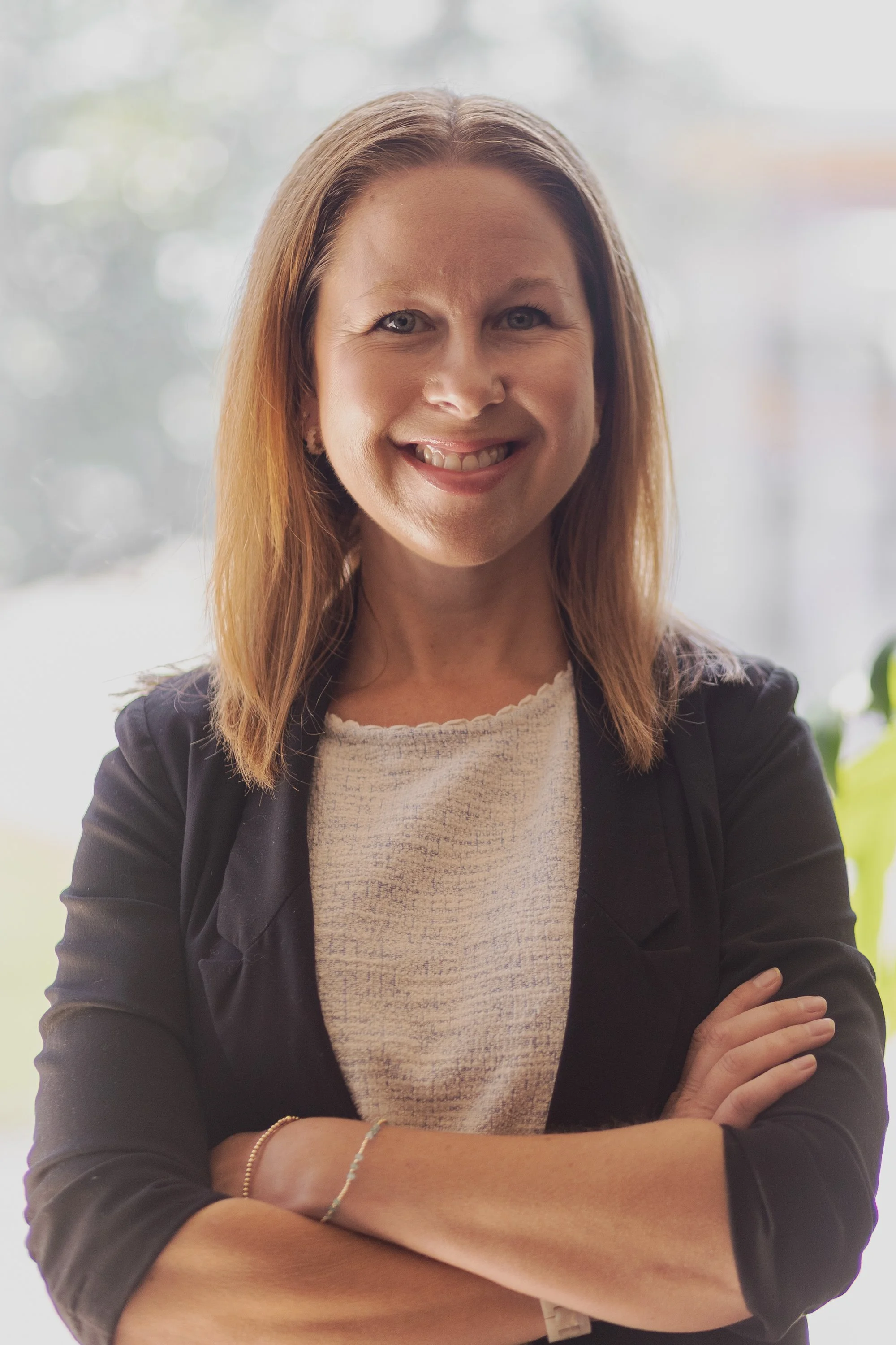 A smiling woman with shoulder-length red hair wearing a black blazer over a white textured top, standing with arms crossed indoors near a window with blurred outdoor background.