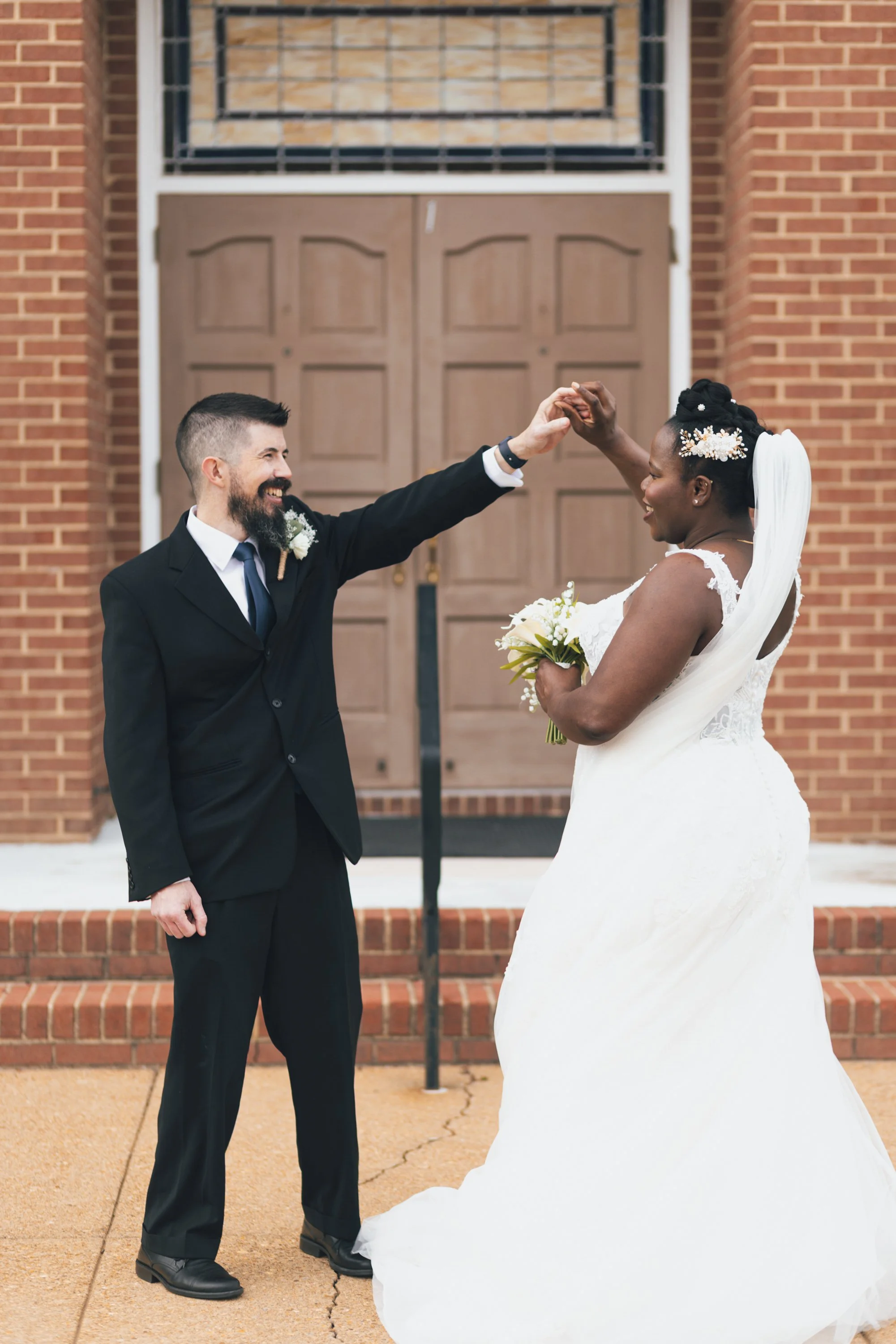 A groom and bride are outside a brick building, holding hands and smiling at each other, dressed in wedding attire. The groom is wearing a black suit and tie, and the bride is in a white wedding dress holding a bouquet of flowers.