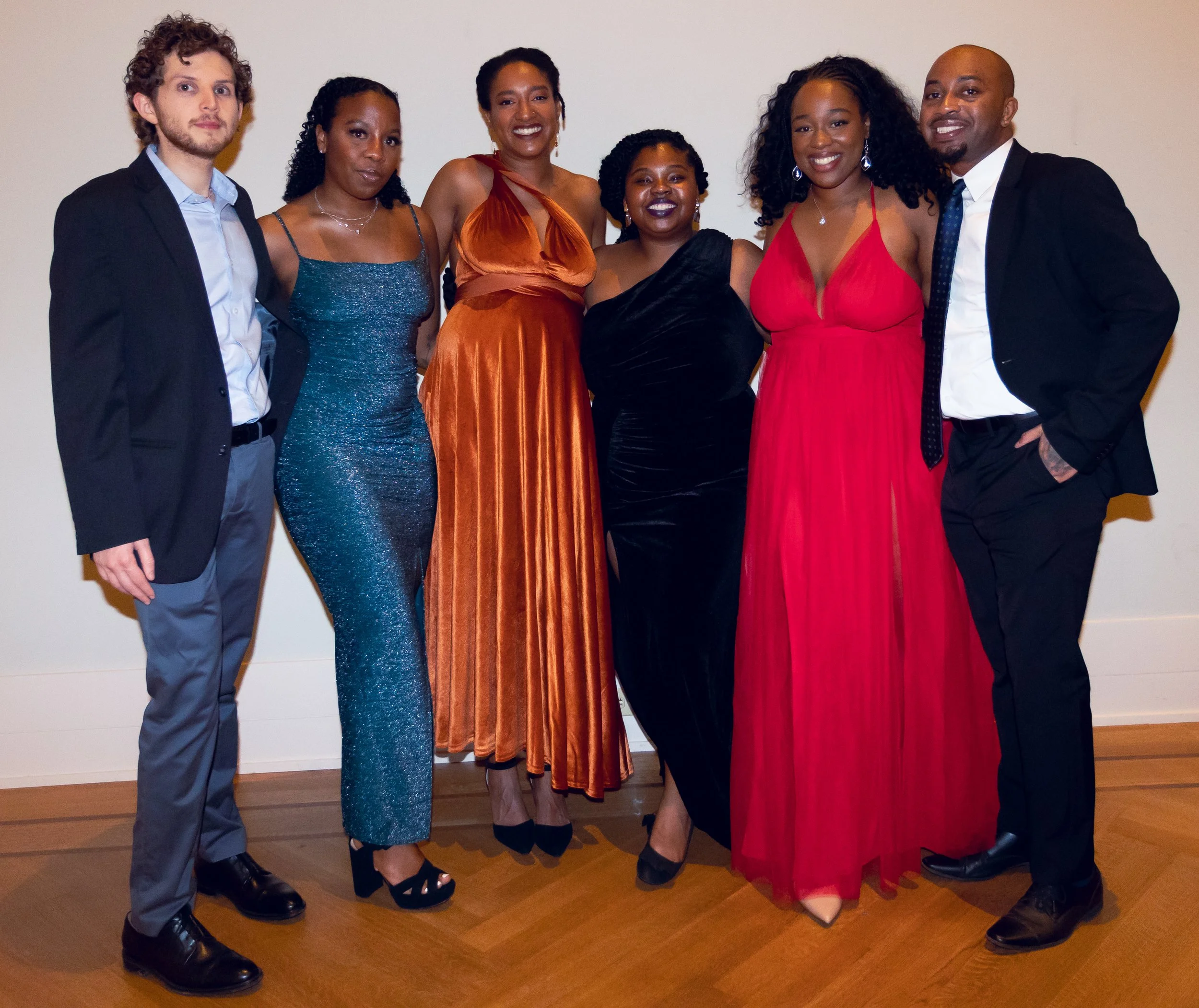 Group of six people dressed in formal attire standing together indoors against a plain wall, smiling and posing for a photo.