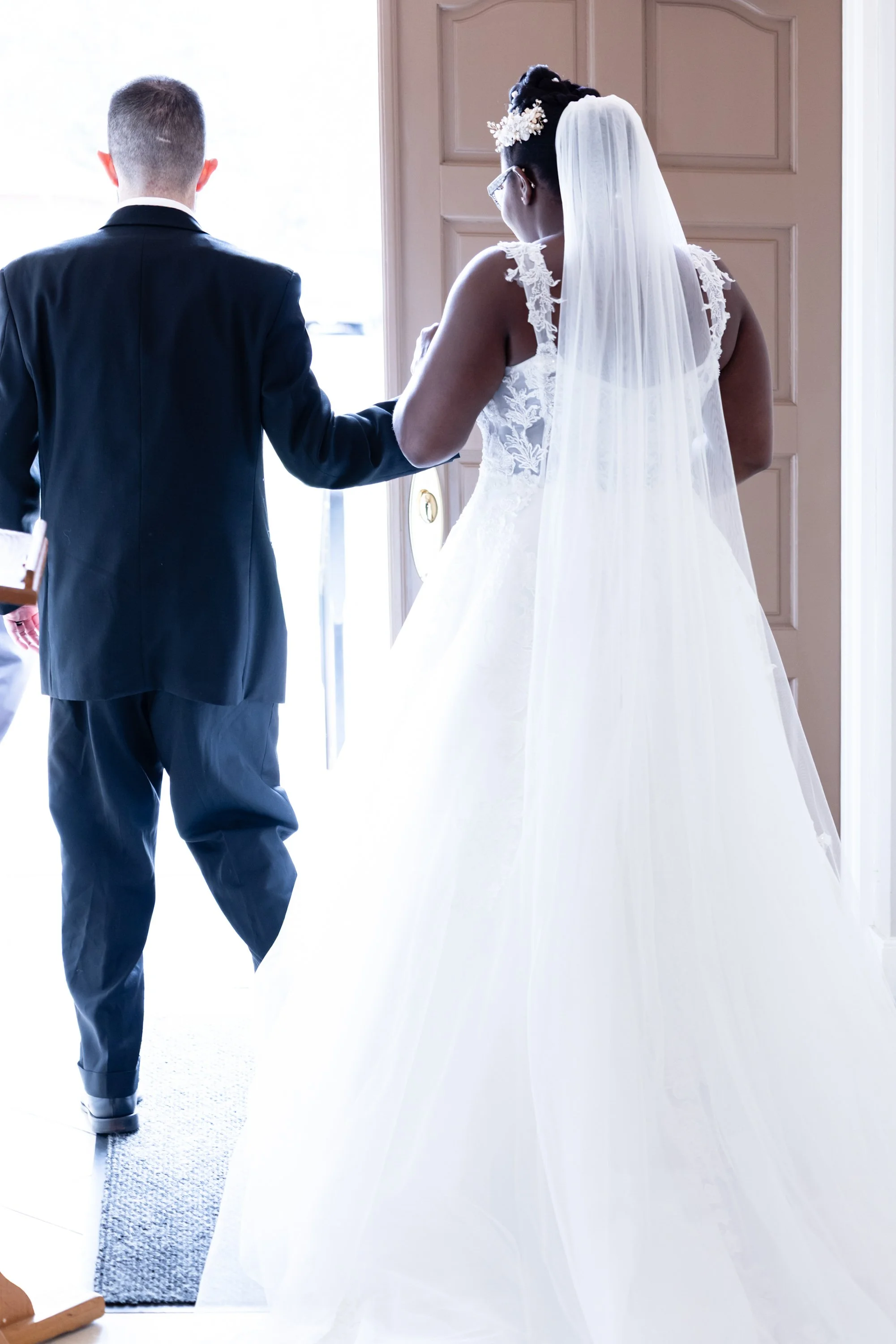 A bride and groom entering a room, the bride in a white lace wedding dress with a long veil and floral hairpiece, the groom in a black tuxedo, holding hands, with natural light coming from outside.