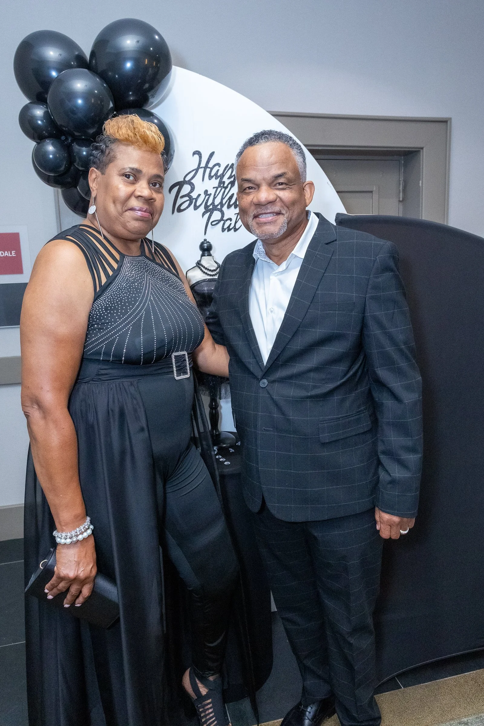 Two people standing in front of a birthday celebration backdrop with black balloons, a white circle with black writing, a black dress, and a man in a suit. The woman is wearing a black dress with rhinestone details, and the man is dressed in a dark c