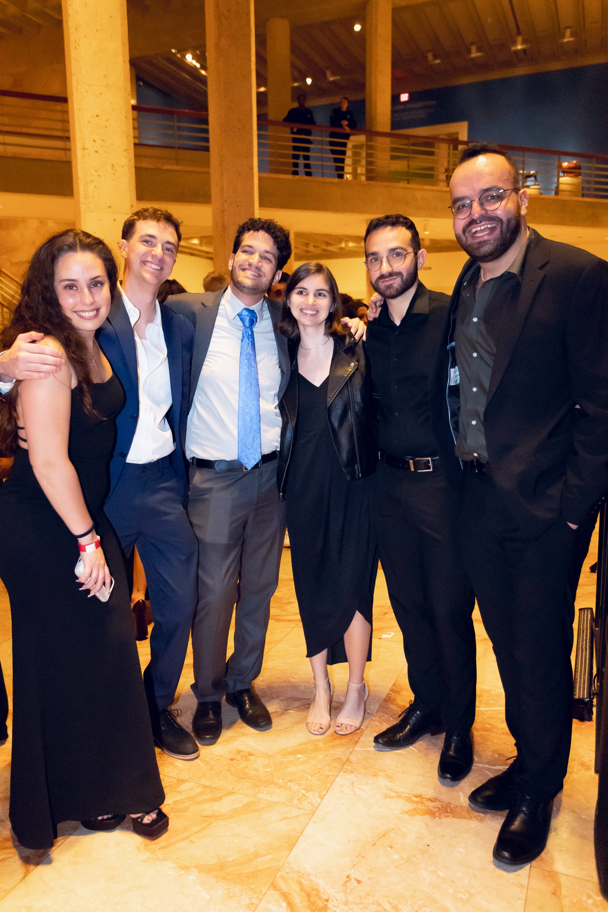 Group of six young adults dressed professionally, smiling and posing together in an indoor event space with wood accents and warm lighting.