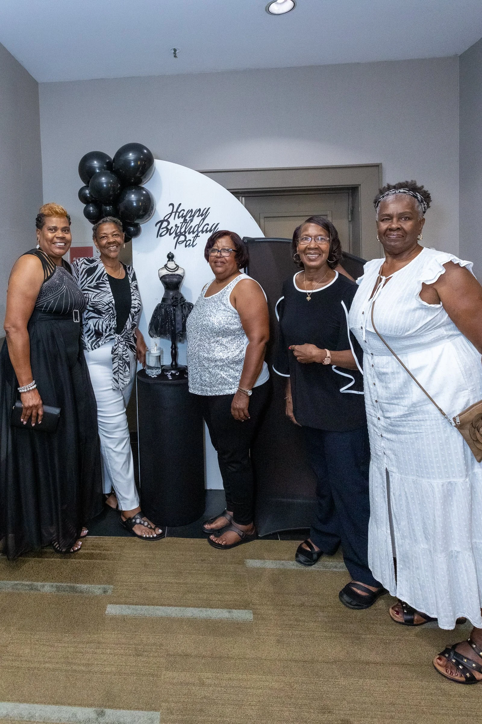 Group of five women standing in front of a birthday celebration display with black balloons and a mannequin dress, smiling for the camera.