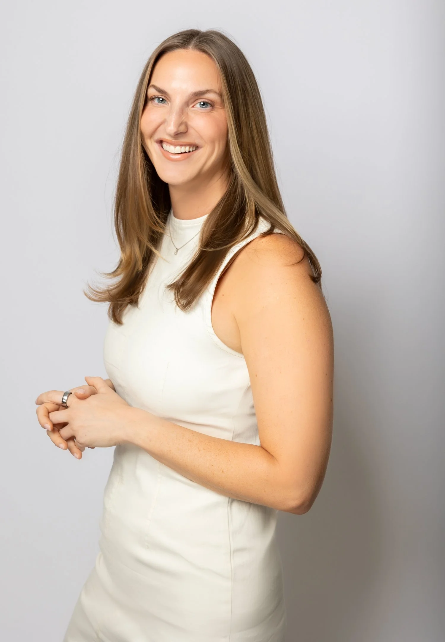 A woman with long, light brown hair and blue eyes smiling, wearing a sleeveless white dress and jewelry, standing against a plain grey background.
