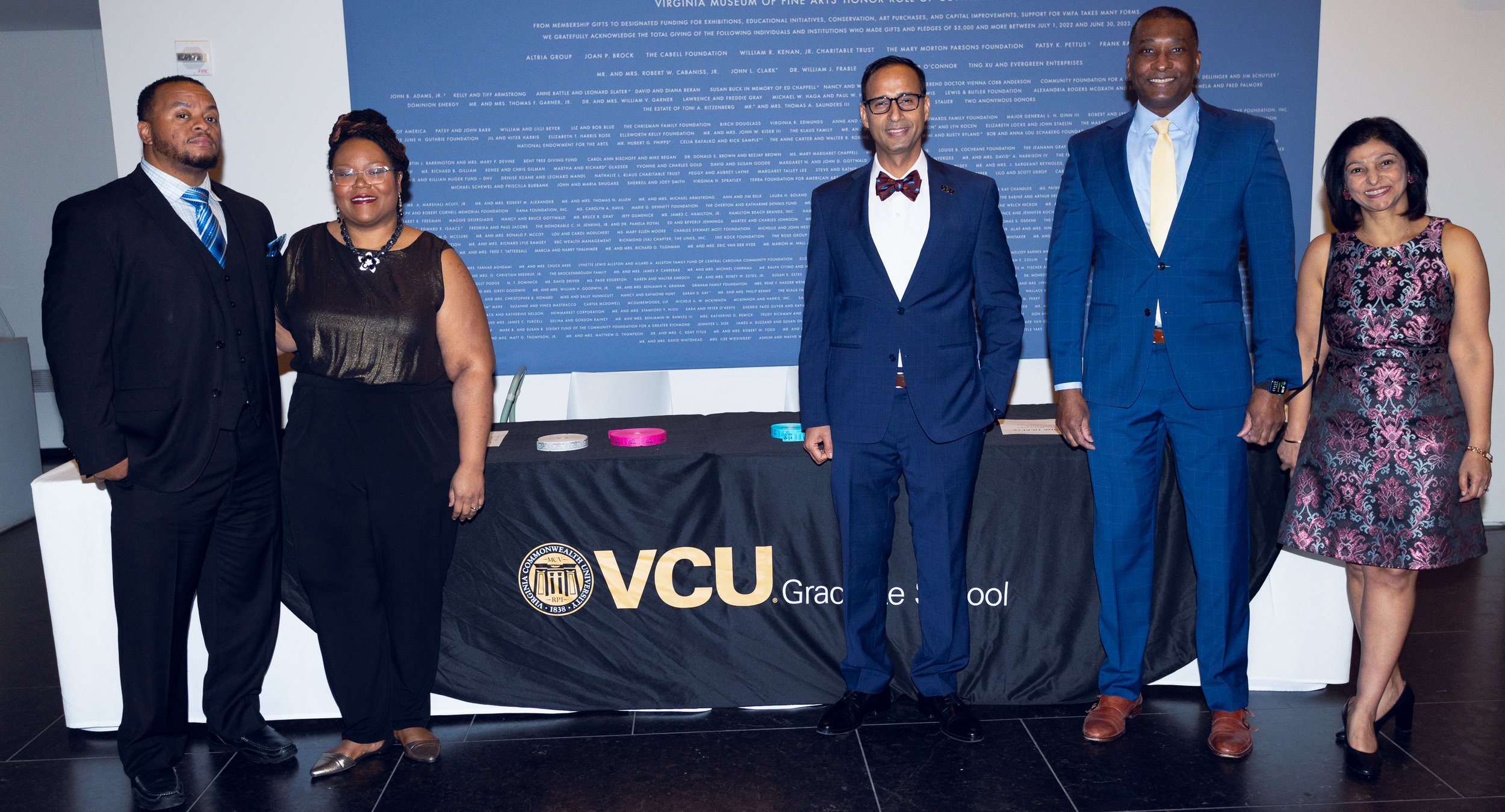 Group of six diverse individuals standing behind a black table with the Virginia Commonwealth University logo, at an event. They are dressed in business attire, posing in front of a blue wall with text.