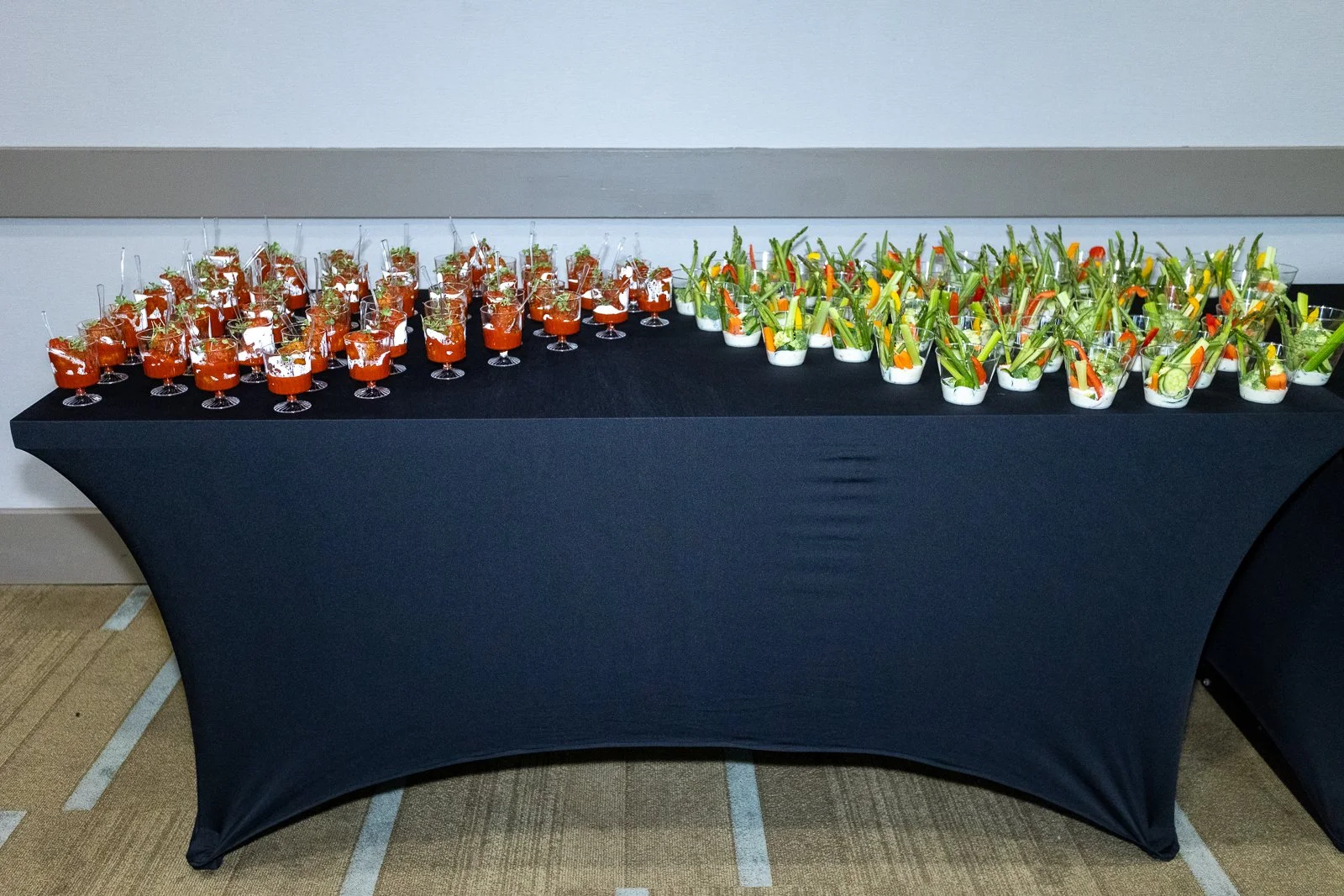 Table with two types of appetizers: on the left, small glasses filled with a red drink, garnished with herbs; on the right, clear cups with vegetable sticks, cucumbers, carrots, celery, and cherry tomatoes, all arranged for a party or event.