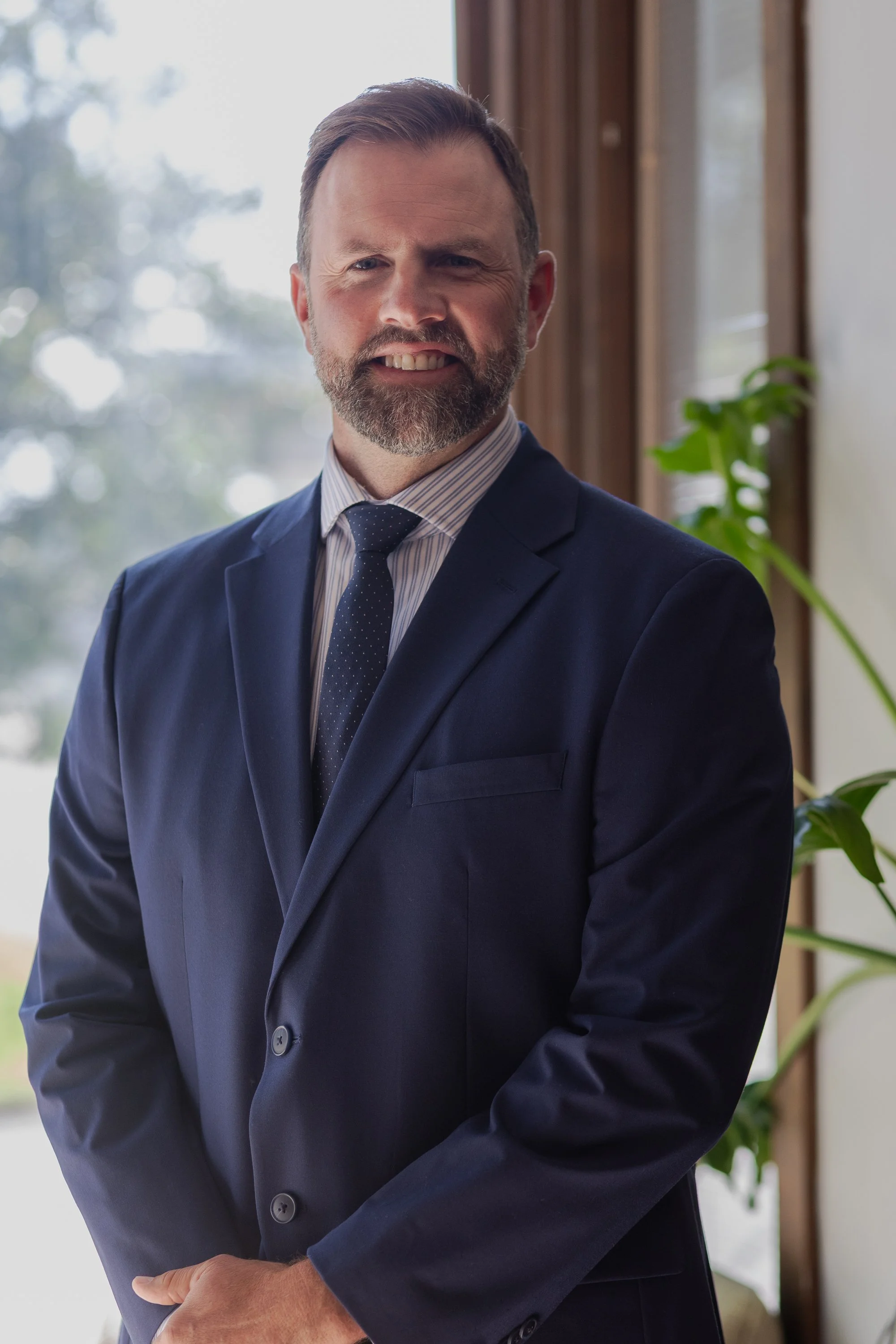 A man with a beard in a blue suit, striped shirt, and dotted tie, standing indoors near a window with natural light and a green plant in the background.