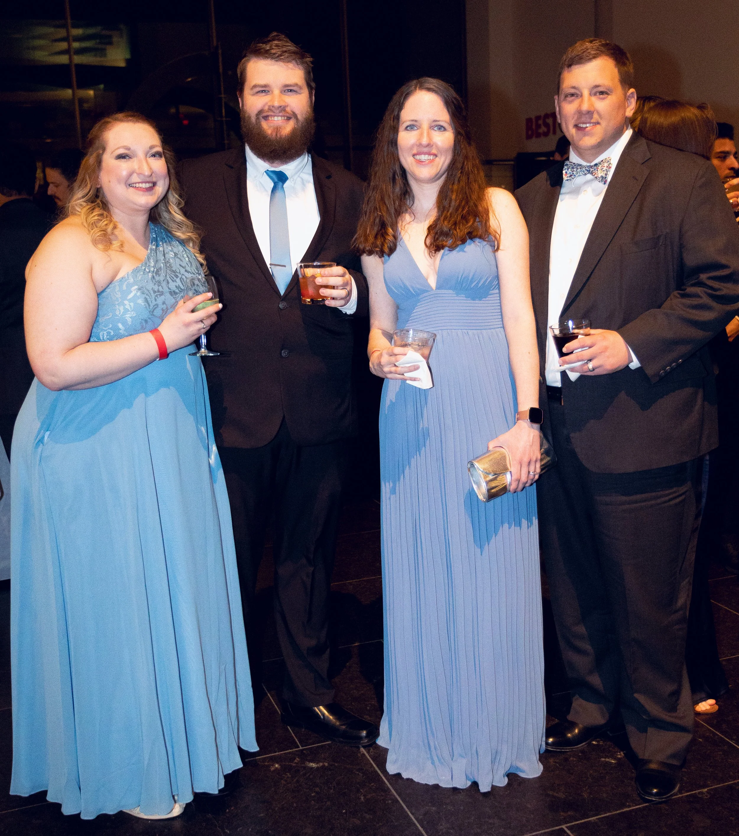 A group of four adults dressed in formal attire at a social event, standing close together and smiling at the camera, with drinks in their hands.