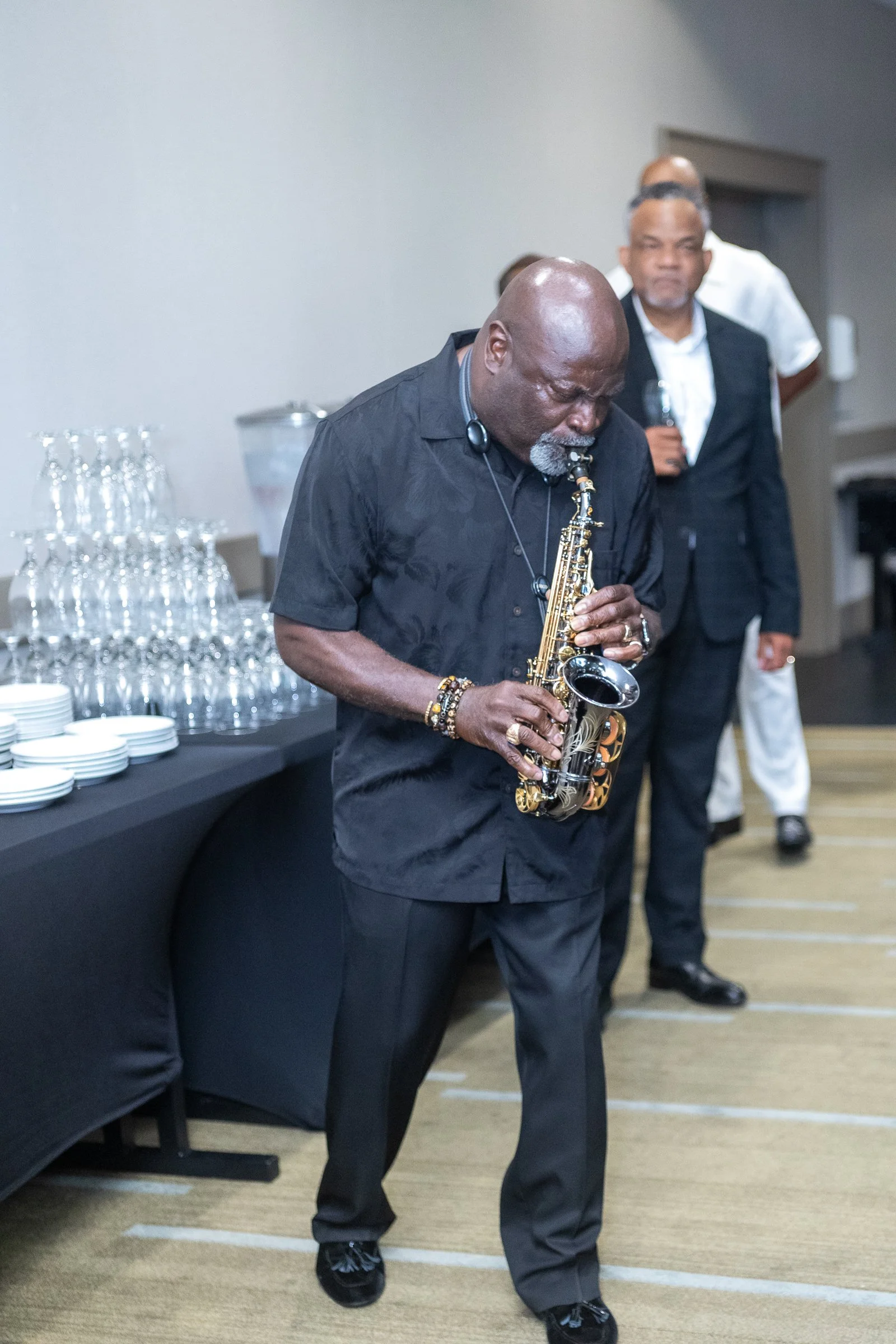 An elderly African American man playing a saxophone at a social event. He is dressed in black, with short gray hair and a beard. In the background, there are other people, and a table with glassware and plates.