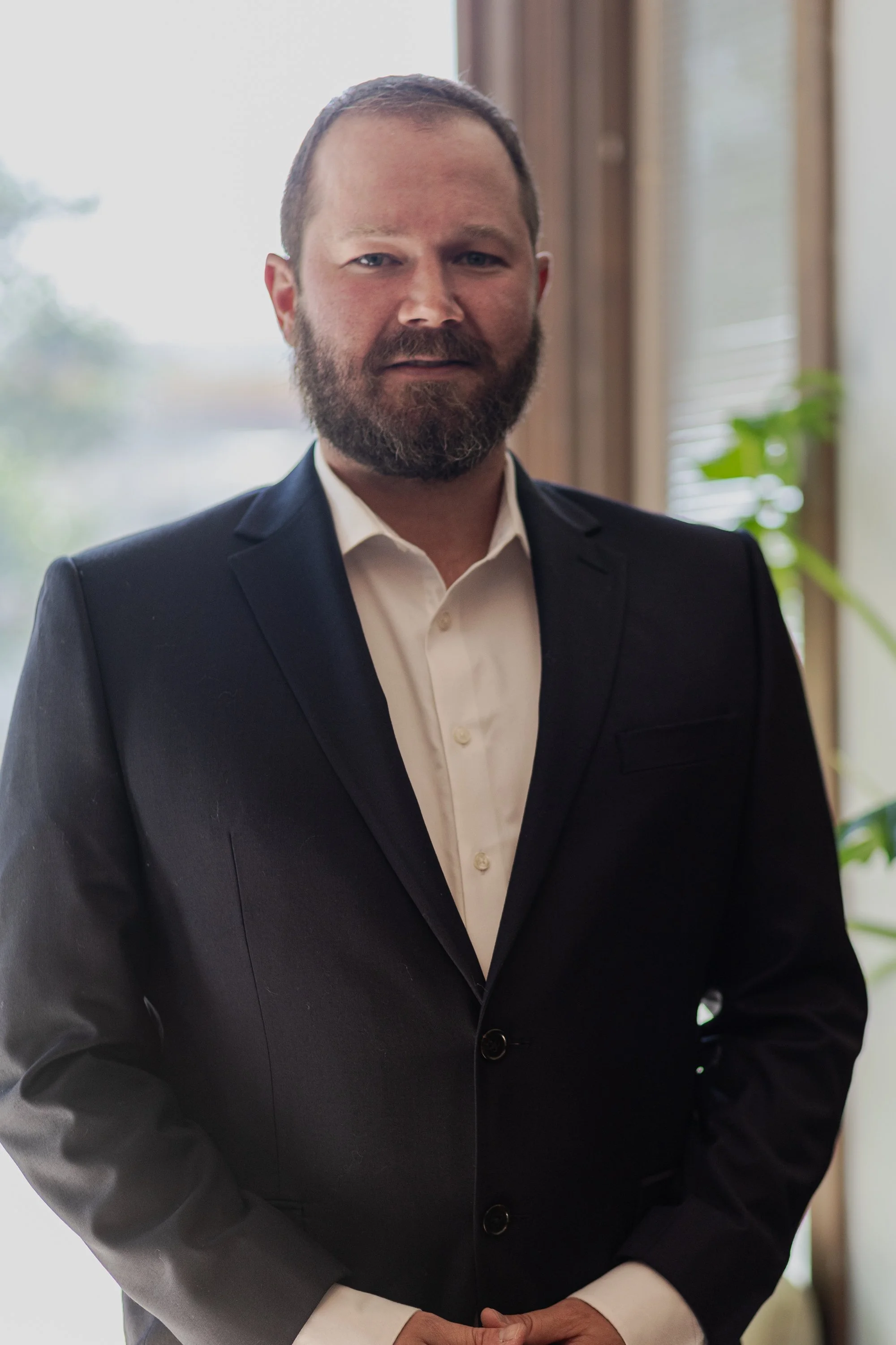 A man in a dark suit with a white shirt standing indoors near a window with blinds, with greenery and natural light in the background.