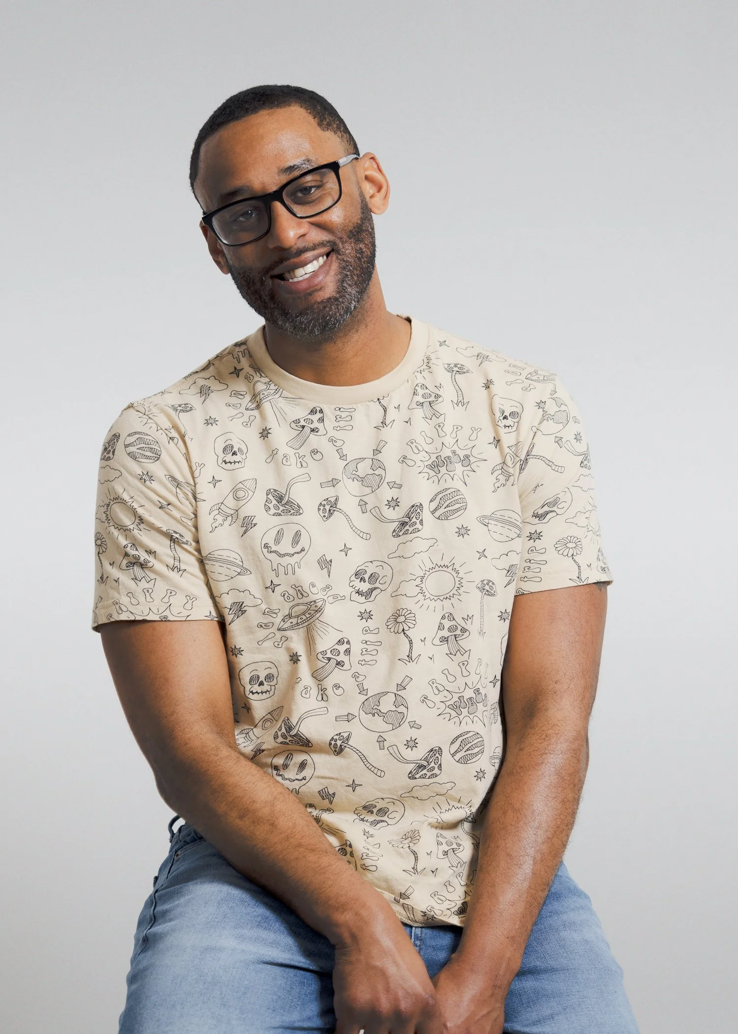 A man with glasses, dark hair, a beard, wearing a beige T-shirt with black doodles of skulls, planets, rockets, and other space-themed drawings, sitting against a plain gray background.