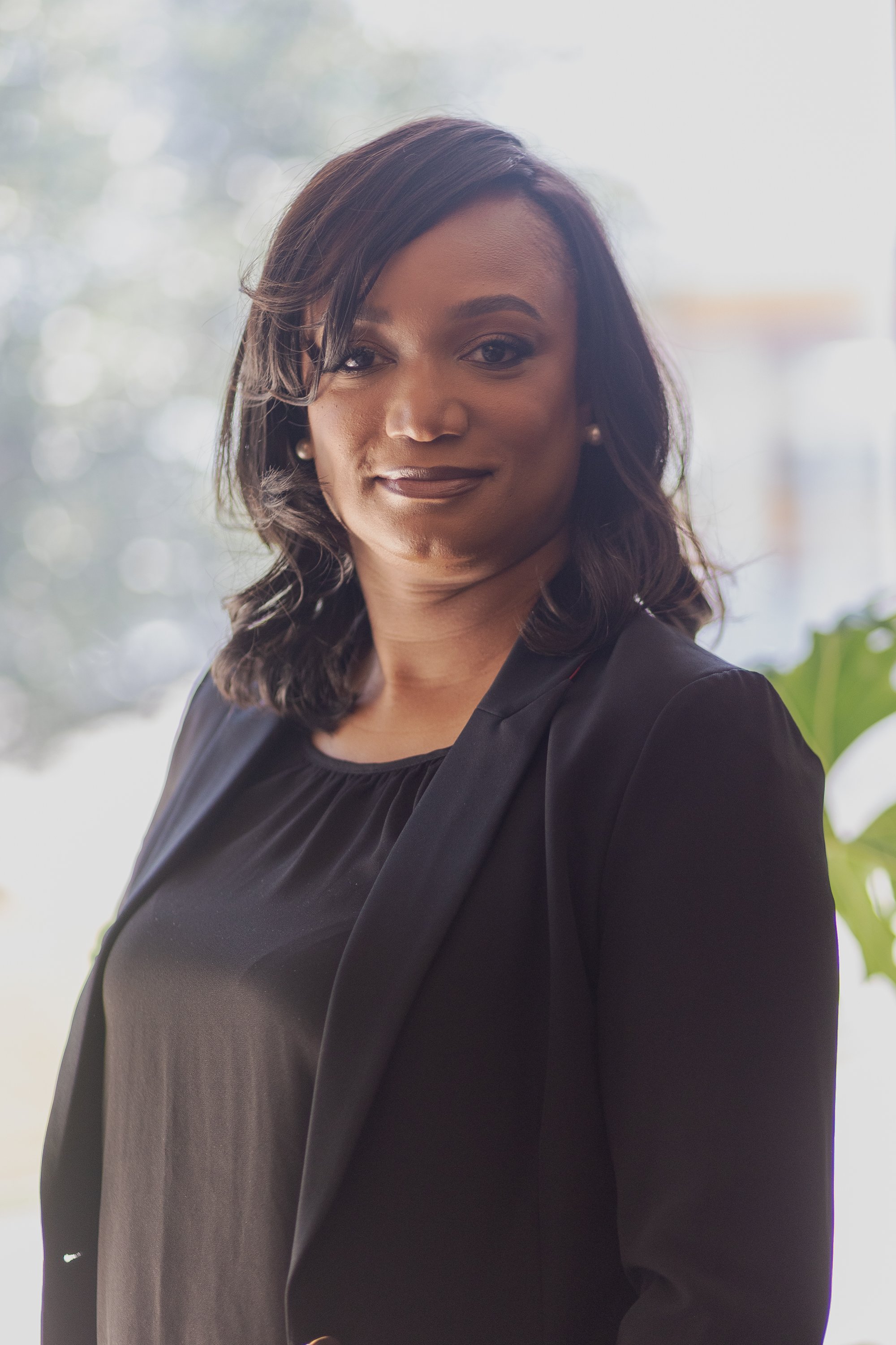 A woman with dark hair and a dark professional outfit smiling indoors with a bright, out-of-focus background.