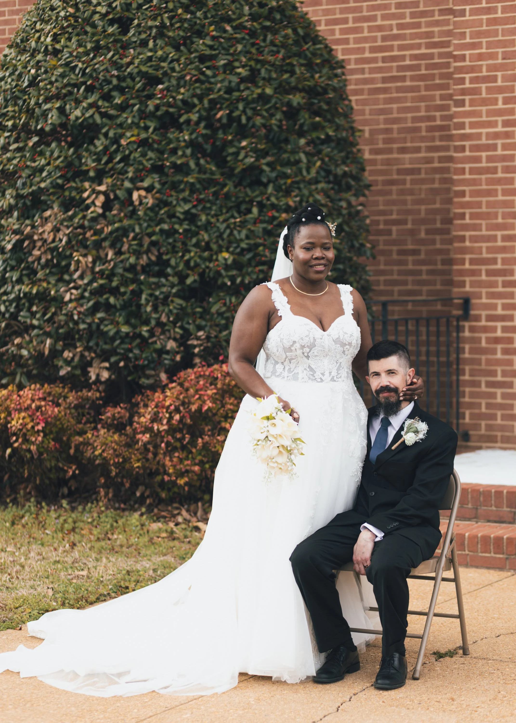 A bride in a white wedding dress standing next to a seated groom in a black tuxedo outside on a brick pathway with a large bush and brick building in the background.