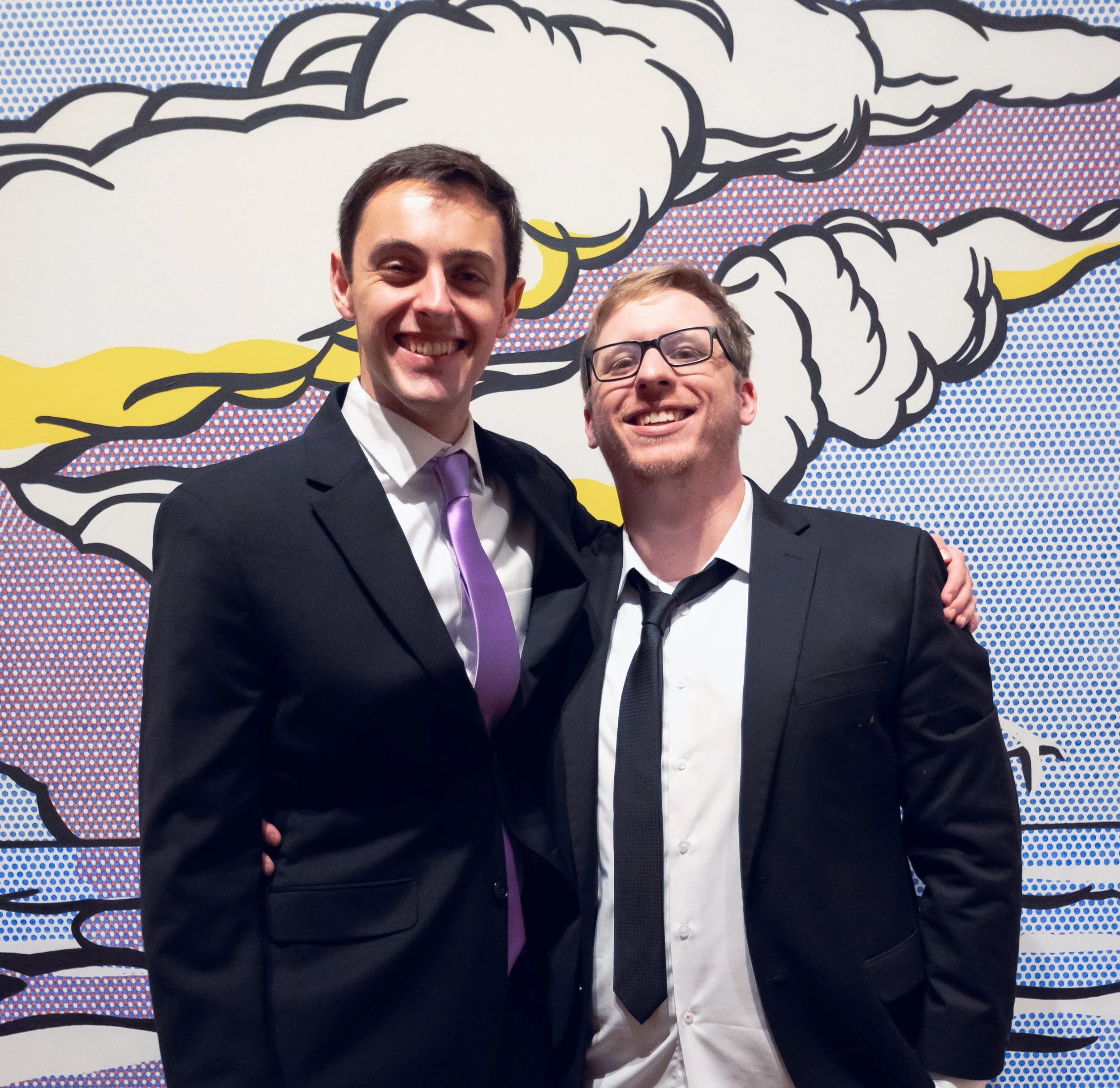 Two men in formal attire standing in front of a colorful pop art-style mural with black outlines and halftone dots, smiling and posing for a photo.