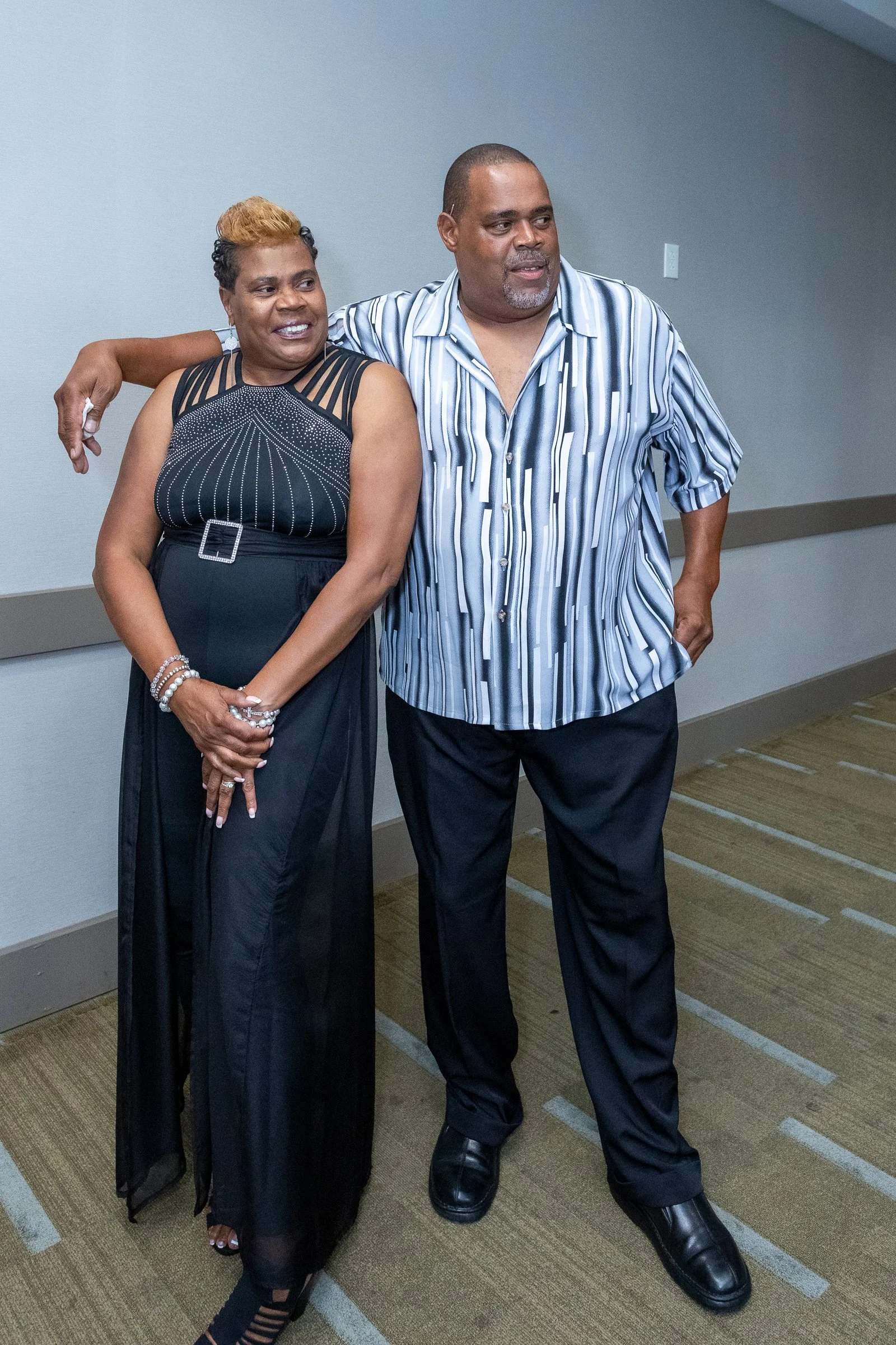 Two people, a woman and a man, standing close together in a hallway. The woman is wearing a black dress with sheer straps and smiling, the man has his arm around her shoulder, wearing a striped shirt and dark pants, looking slightly to the side.