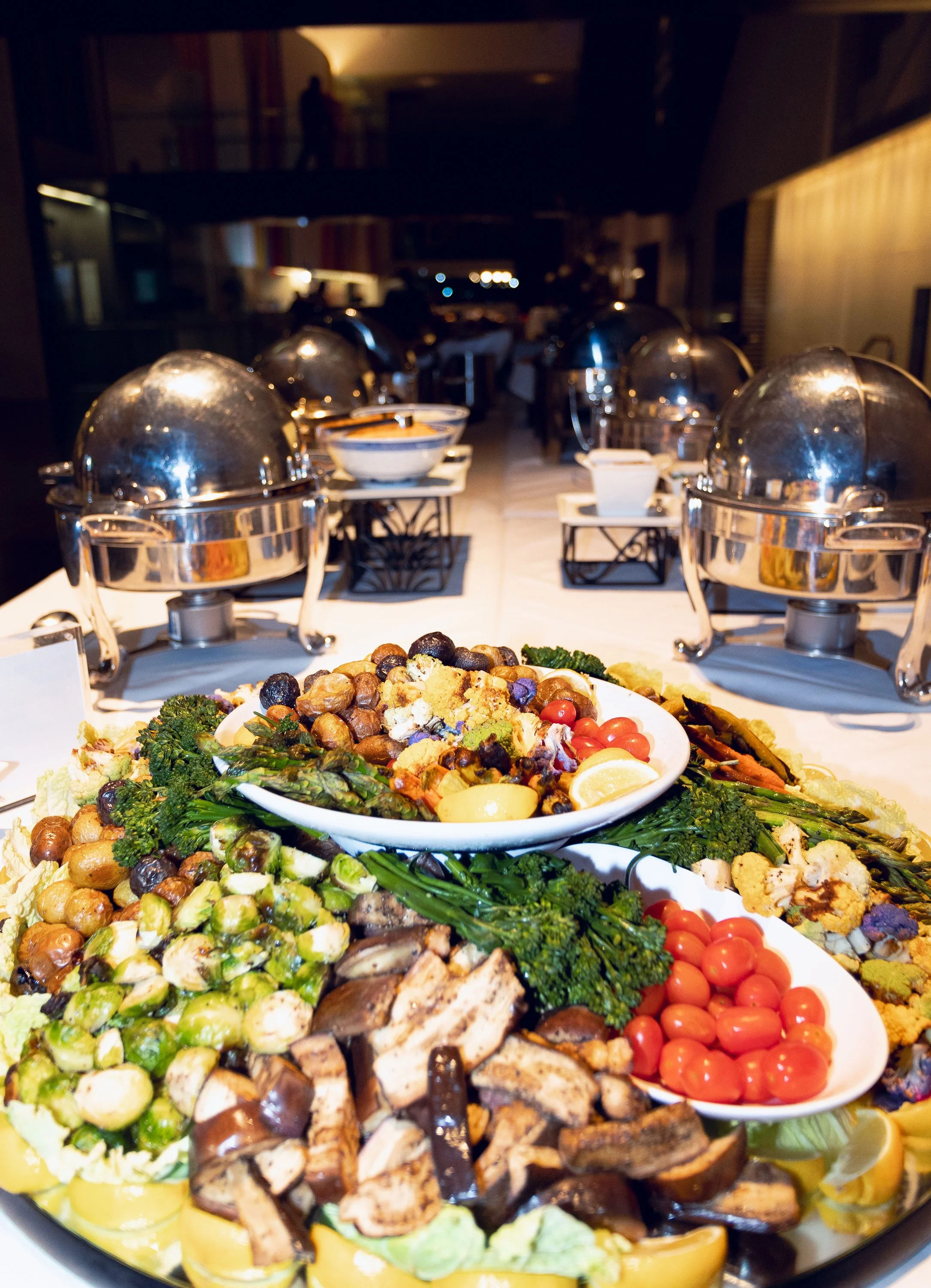 A buffet table with various dishes, including a large platter of salads, vegetables, and grilled meats, with stainless steel chafing dishes in the background in a dimly lit restaurant.