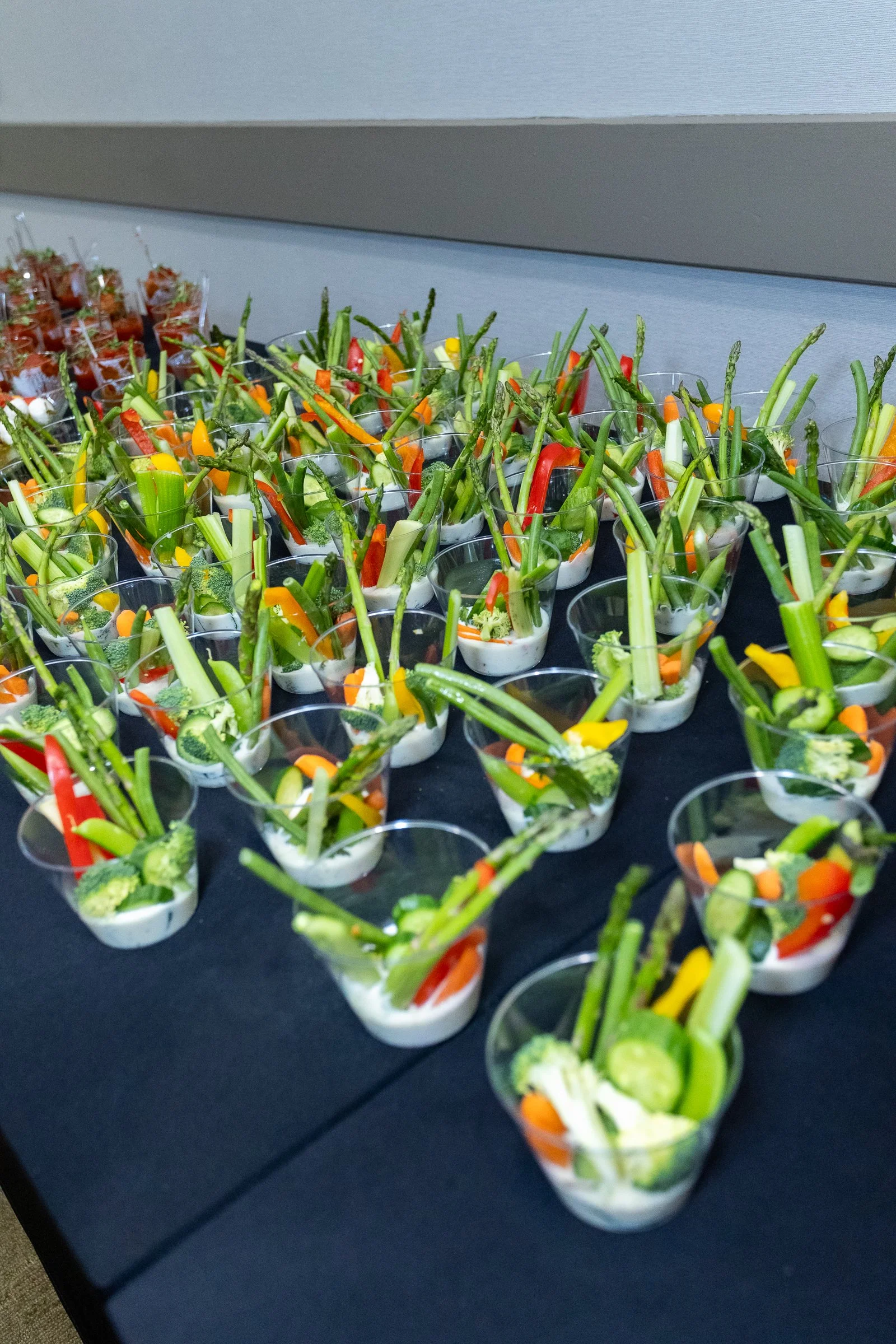 Assorted vegetable appetizers in small clear cups on a black table, featuring sliced cucumbers, bell peppers, broccoli, and carrots.