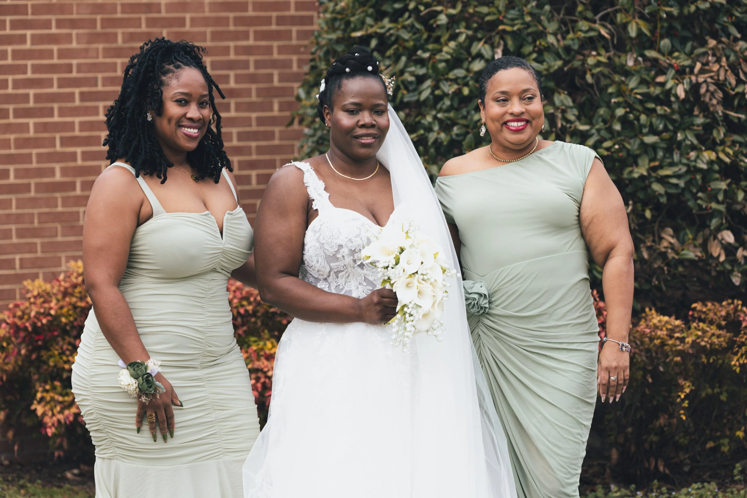 Three women standing outdoors, one dressed as a bride in a white wedding gown holding a bouquet, and two women dressed in light green dresses, posing together in front of a brick wall and green foliage.