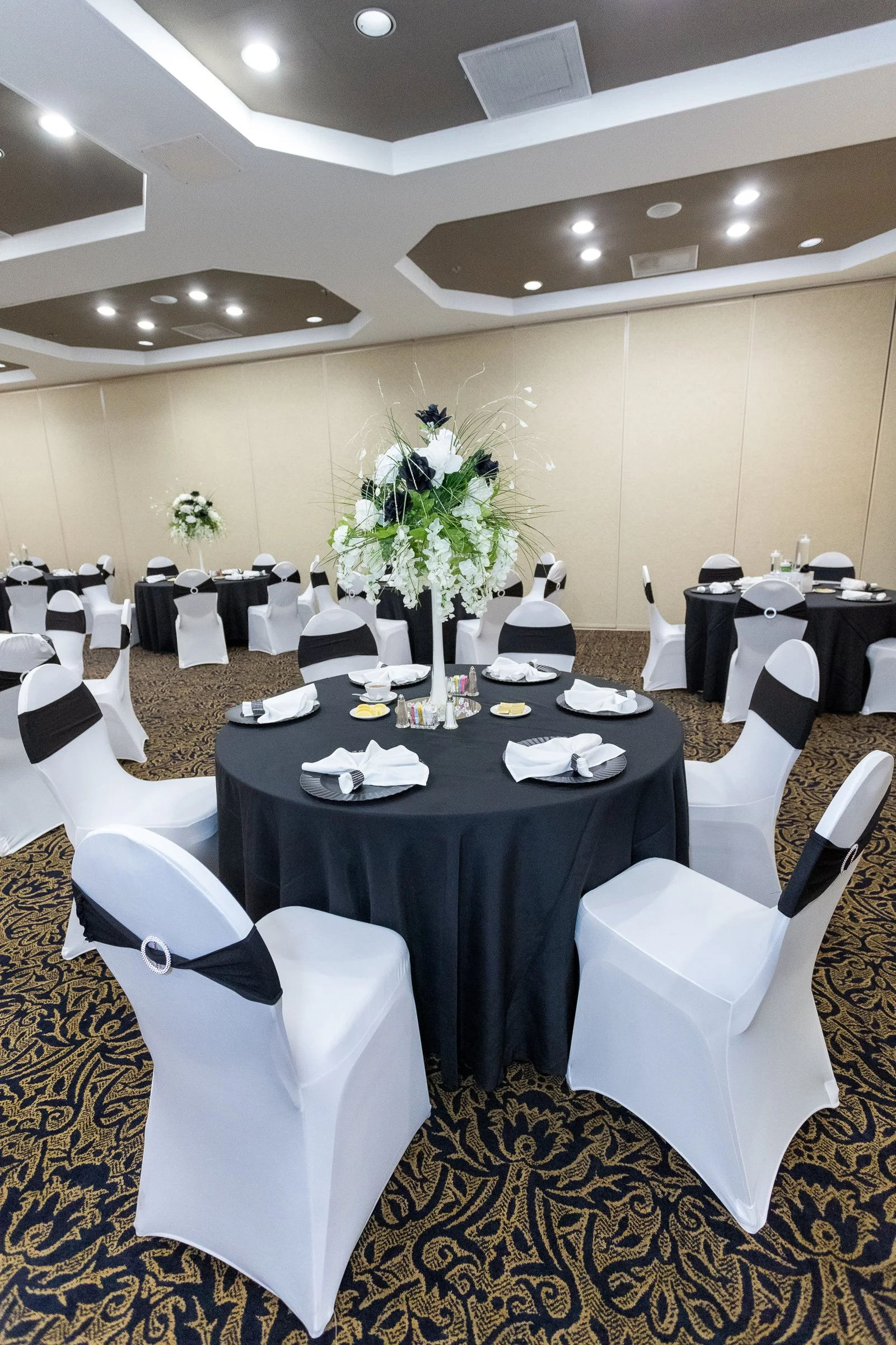 Banquet hall decorated for an event with tables covered in black tablecloths and white chairs accented with black sashes. Centerpieces feature tall floral arrangements with white and black flowers.