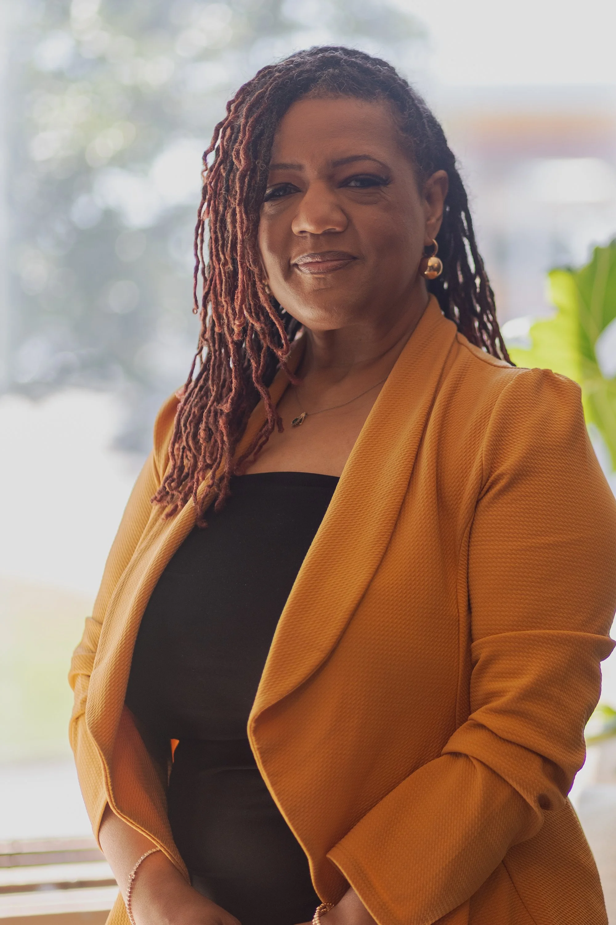 A woman with dreadlocks wearing a mustard colored blazer over a black top, accessorized with gold earrings and necklace, standing outdoors with a blurred background of trees and sky.